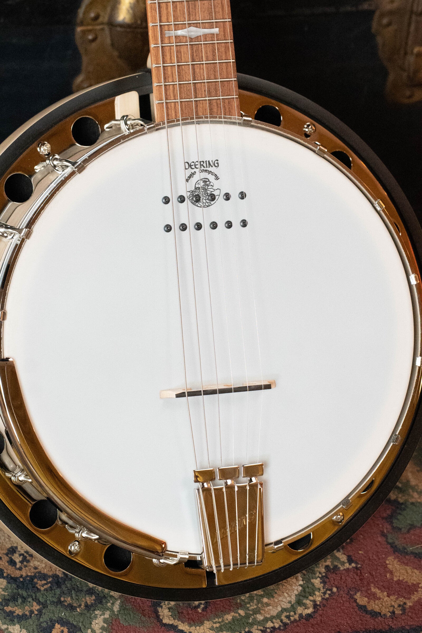 A close-up of the Deering Banjos Goodtime Six-R 6-String Resonator Banjo with Kavanjo Pickup, highlights its white drumhead, metal strings, wooden bridge, and gold hardware as it rests on a colorful patterned carpet.