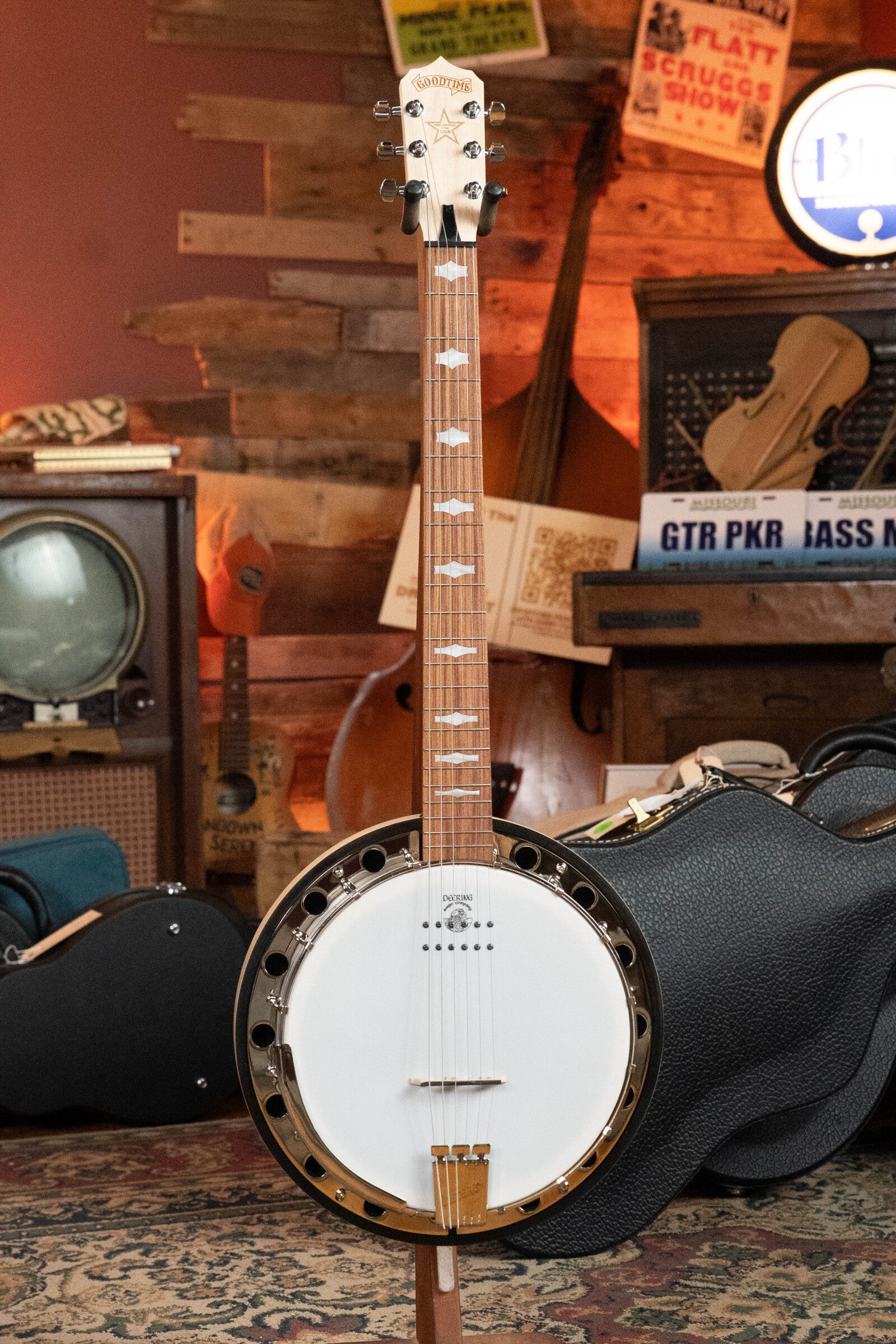 The Deering Goodtime Six-R 6-String Resonator Banjo w/Kavanjo Pickup by Deering Banjos, with a light neck, white drumhead, and gold accents, stands upright on a patterned rug in a music-themed room filled with vintage decor and instruments.