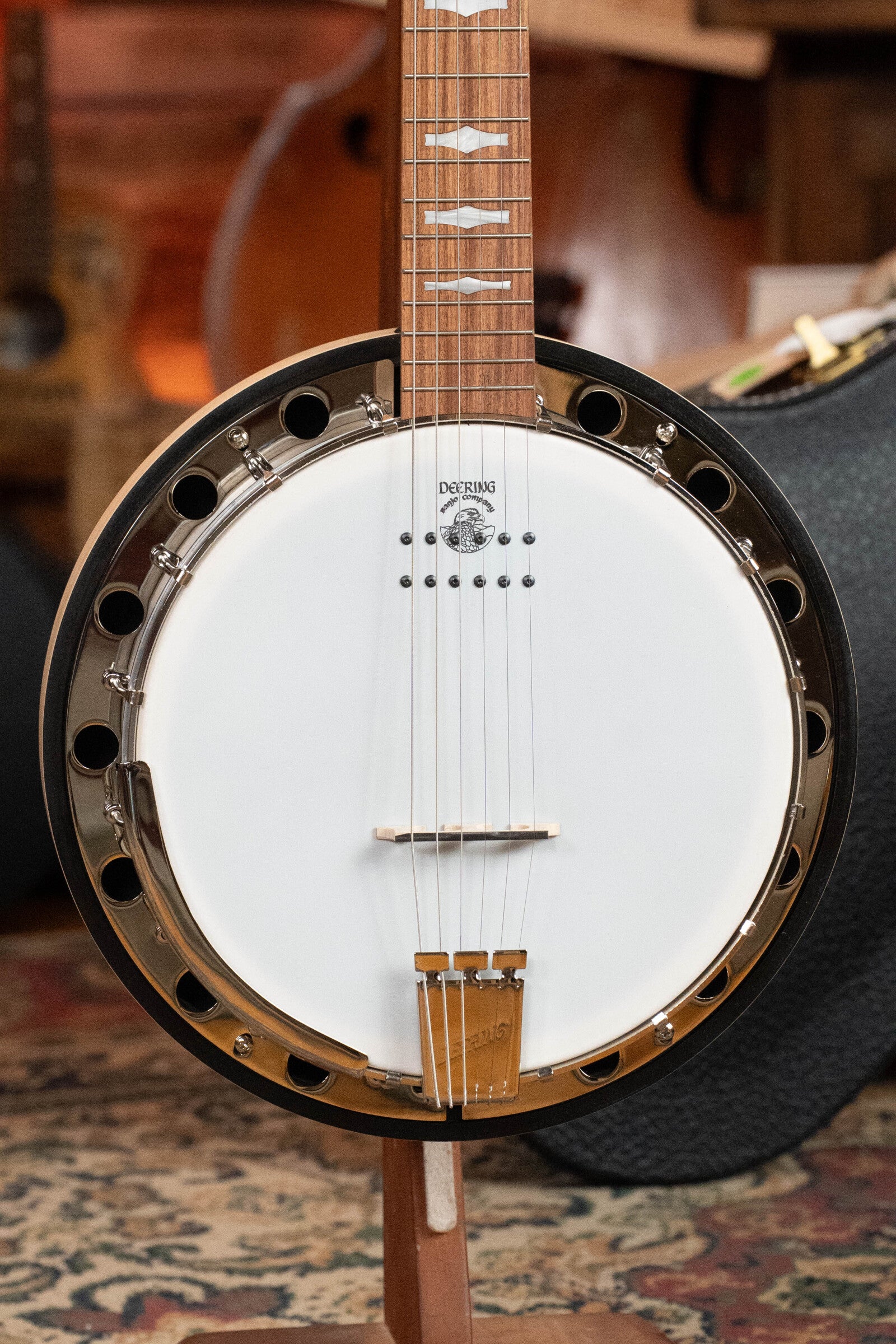 A close-up of the Deering Goodtime Six-R 6-String Resonator Banjo w/Kavanjo Pickup by Deering Banjos stands upright on a patterned rug, highlighting its strings, circular body, and metallic rim, with blurred musical instruments in the background.