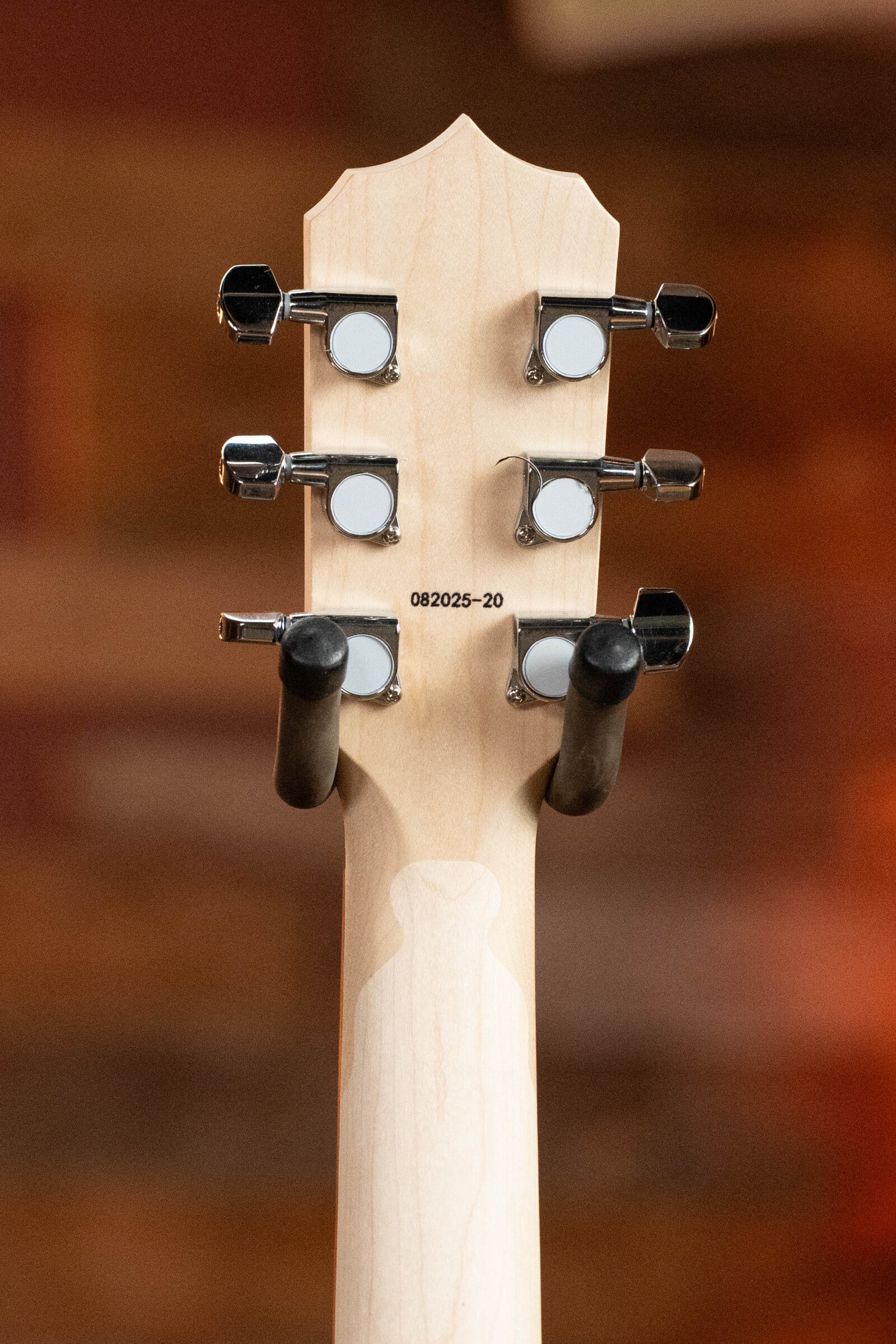 Close-up of the Deering Banjos Goodtime Six-R 6-string resonator banjo w/Kavanjo pickup (#2520) headstock, showing six tuning pegs and serial number, mounted on a stand against a blurred brown background.
