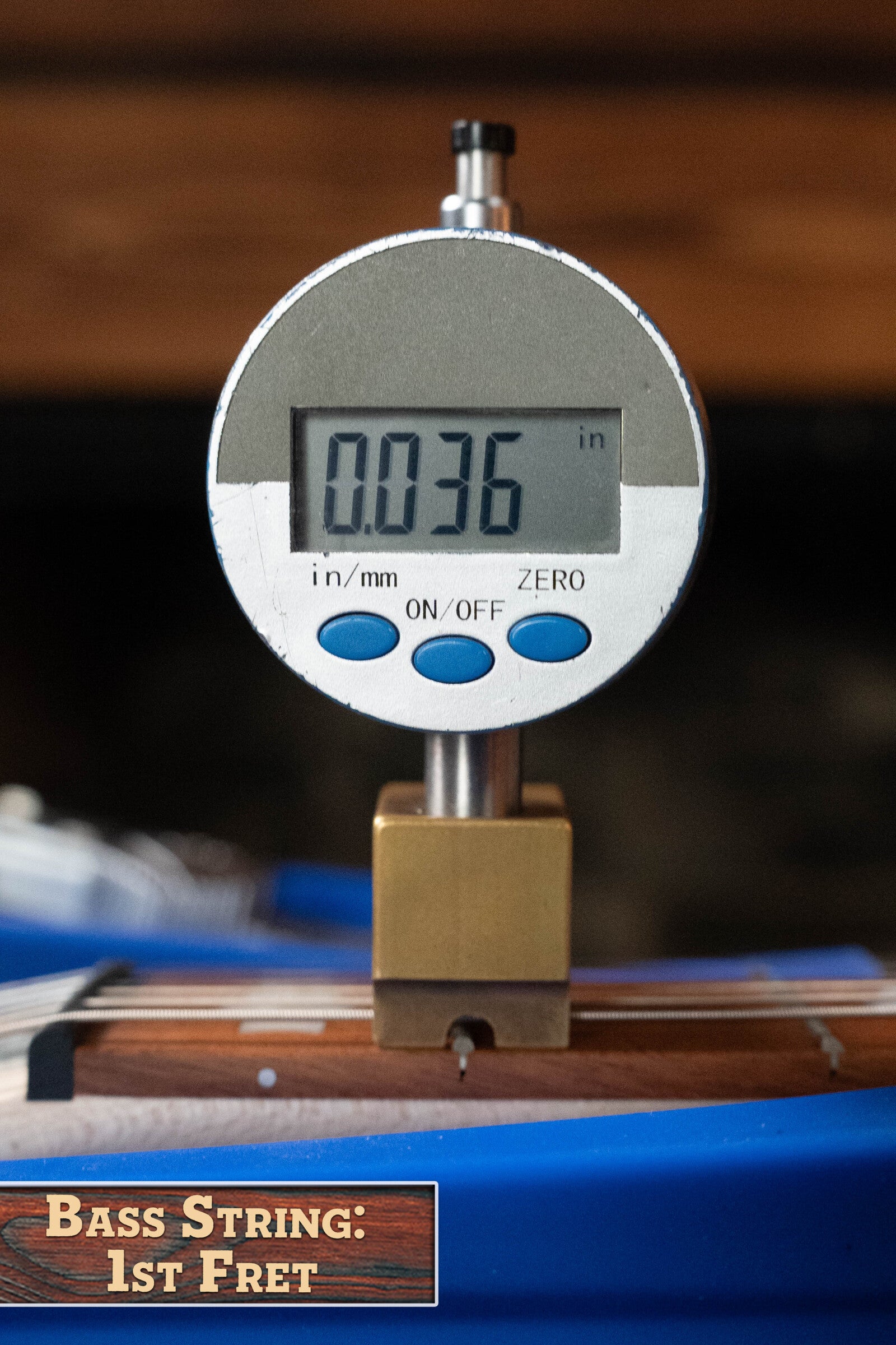 A Deering Goodtime Six-R 6-String Resonator Banjo w/Kavanjo Pickup by Deering Banjos shows a digital caliper reading of 0.036 inches on the bass string at the 1st fret. A wooden label reads: Bass String: 1st Fret.