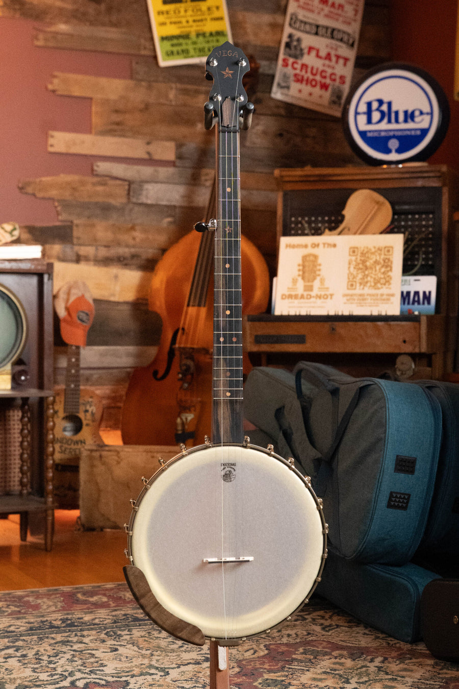 A Deering Vega Vintage Star 5-String Openback Banjo w/Spikes (#BB66) by Deering Banjos stands upright on a patterned rug in a cozy room, surrounded by vintage instruments, posters, and a teal armchair, radiating vintage charm.