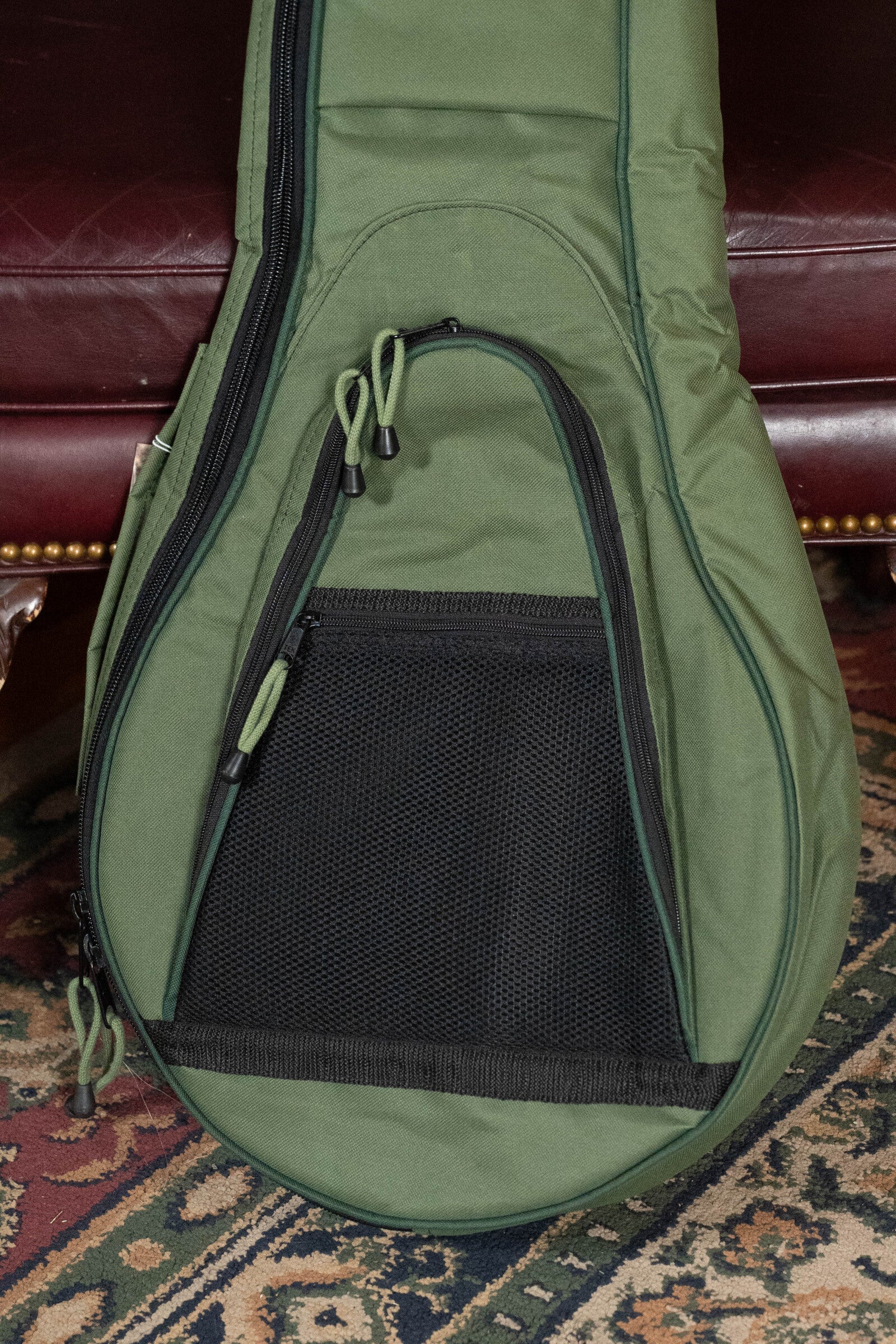 A close-up of the Deering Vintage Eagle Tenor Banjo Gigbag in green by Deering Banjos, featuring a zippered pocket and mesh panel, shown on a patterned rug near a maroon leather couch with brass studs.