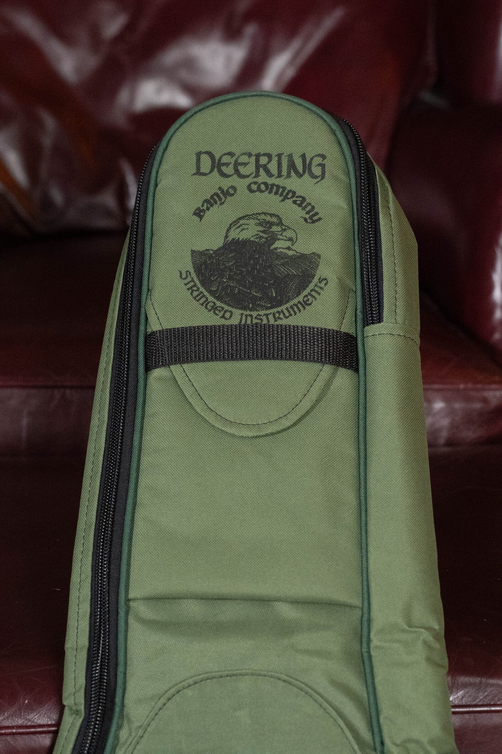 A Deering Vintage Eagle Tenor Banjo Gigbag in green, with black eagle logo and Deering Banjos printed on it, sits on a maroon leather couch.