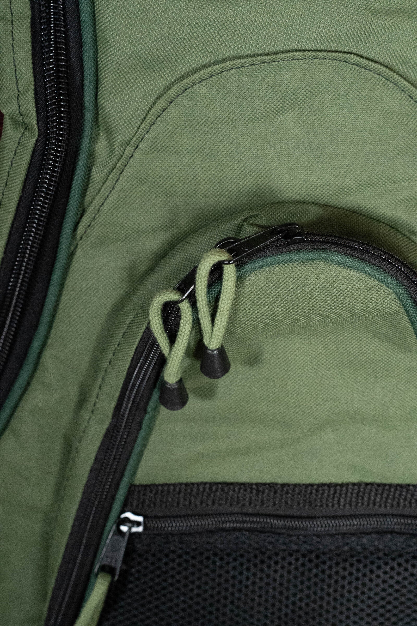 Close-up of the Deering Vintage Eagle Tenor Banjo Gigbag - Green by Deering Banjos, featuring two zippers with green fabric pulls and black plastic ends, a curved zipper pocket, and part of a black mesh pocket at the bottom.