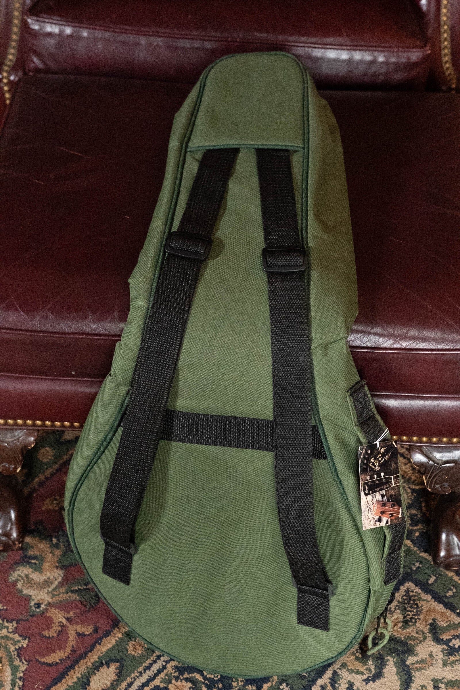 A Deering Banjos Vintage Eagle Tenor Banjo Gigbag in green with black straps rests on a maroon leather chair with brass studs, partly covering a patterned carpet. A tag is attached to the gig bag.