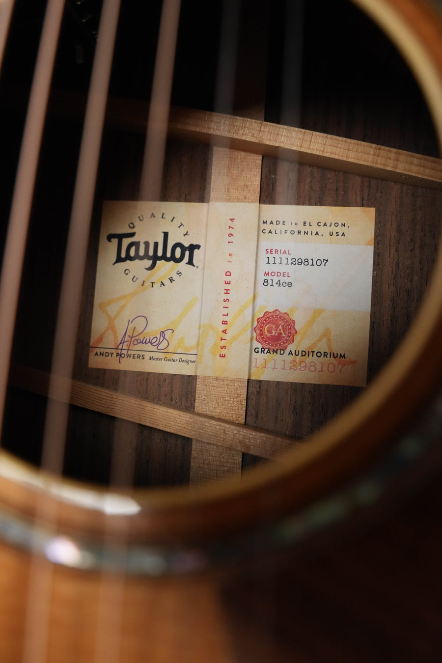 Close-up of a Taylor Guitars *Dread-Not Certified Used* 2018 814ce Grand Auditorium guitar label (#9810) through the strings, showing serial number, V-Class bracing, and signatures on Sitka Spruce/Indian Rosewood grain.