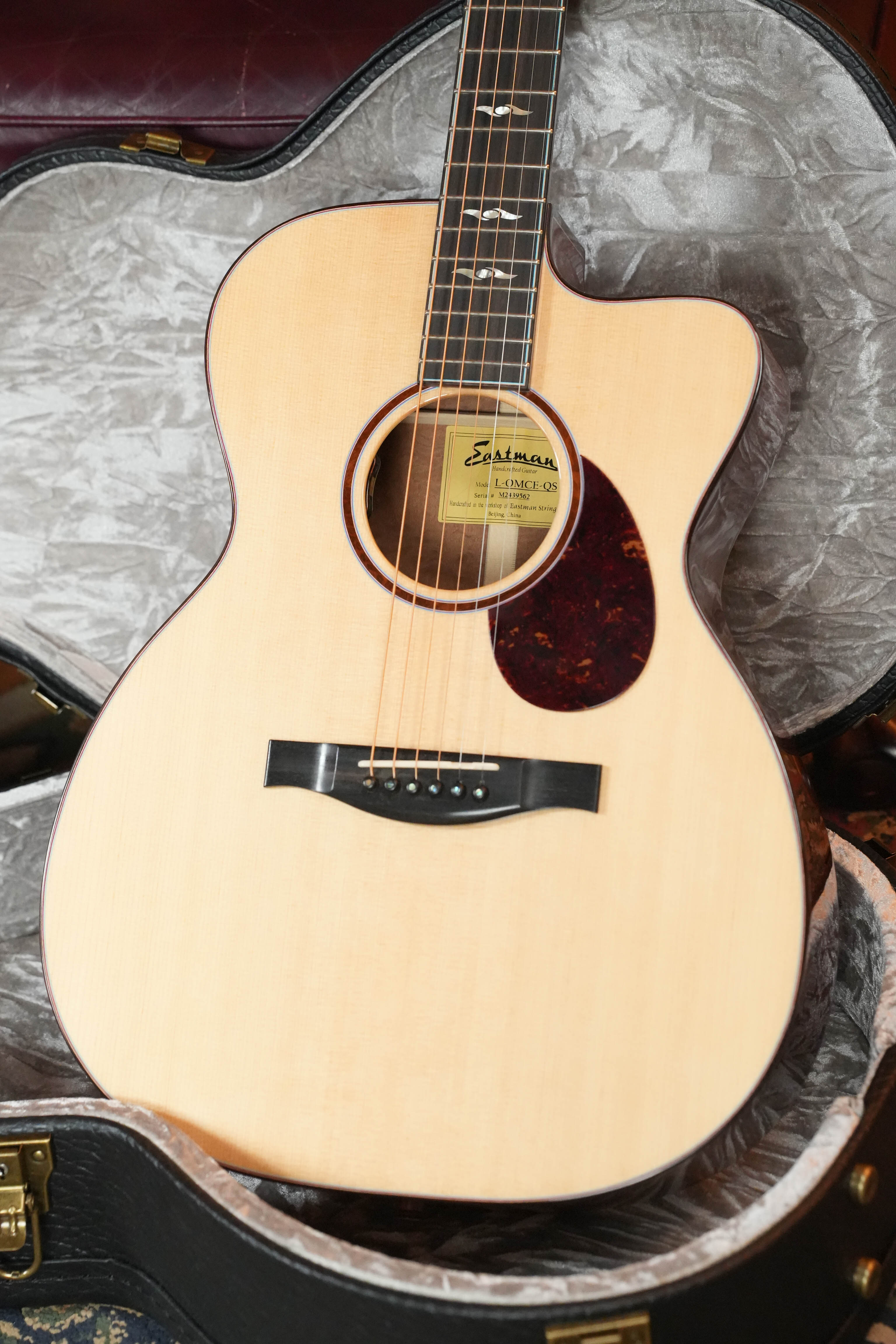 A 2024 Eastman Guitars L-OMCE-QS Orchestra Model acoustic-electric guitar with solid Engelmann spruce top, solid quilted sapele back and sides, and LR Baggs VTC rests in an open hard case with gray plush lining. Certified used by Dread-Not.