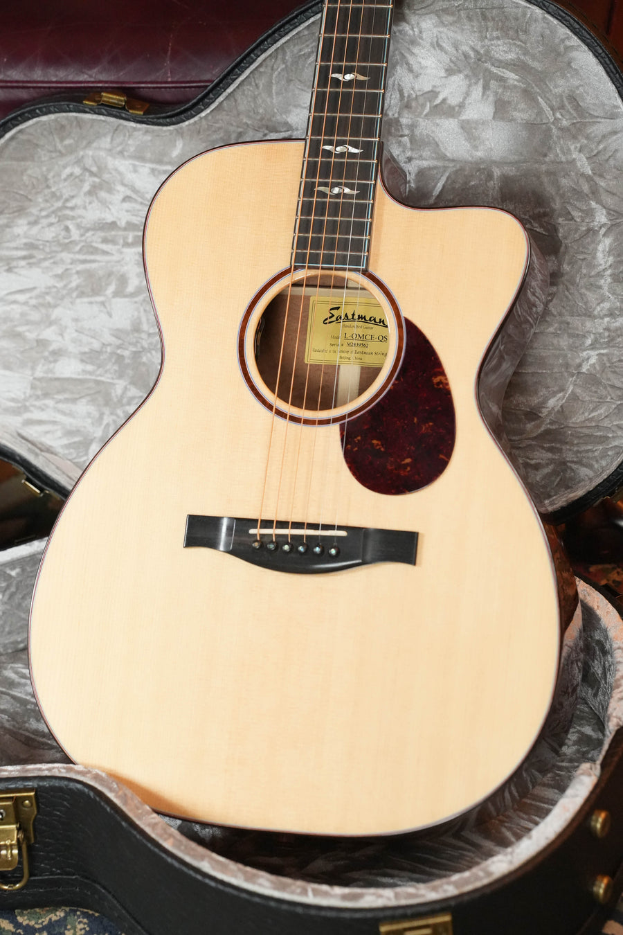 A 2024 Eastman Guitars L-OMCE-QS Orchestra Model acoustic-electric guitar with solid Engelmann spruce top, solid quilted sapele back and sides, and LR Baggs VTC rests in an open hard case with gray plush lining. Certified used by Dread-Not.