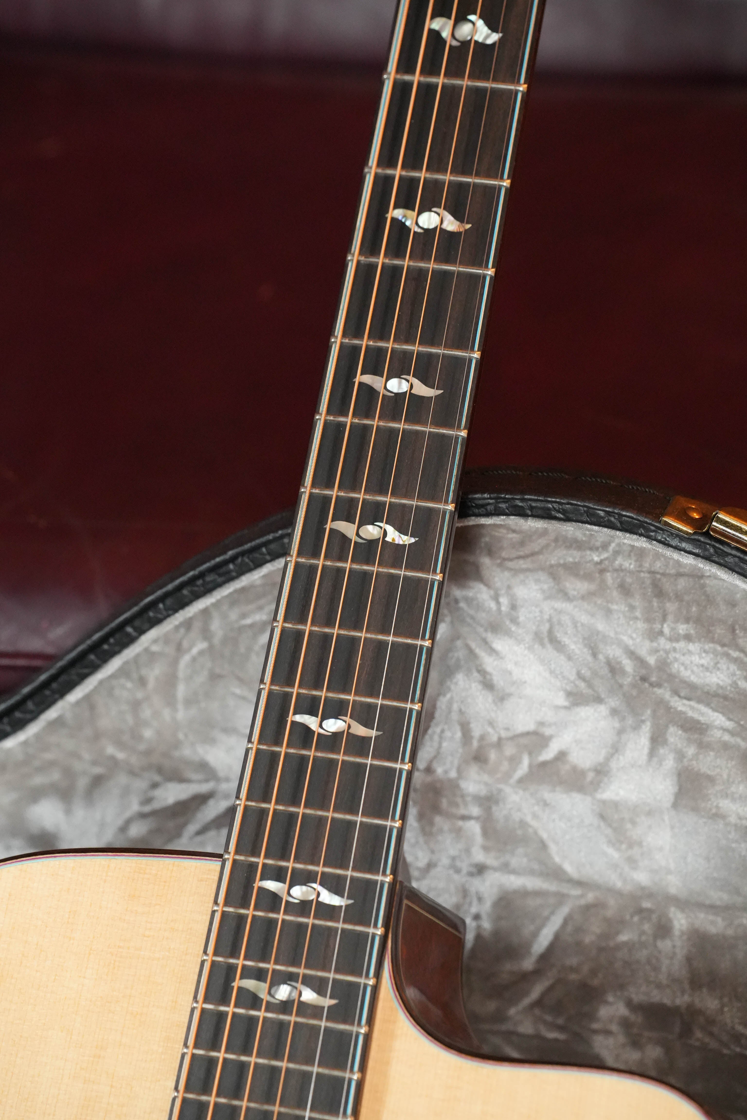 Close-up of a *Dread-Not Certified Used* 2024 Eastman L-OMCE-QS acoustic-electric guitar’s neck and fretboard with dot inlays, shown in an open gray-lined case against a dark red background.
