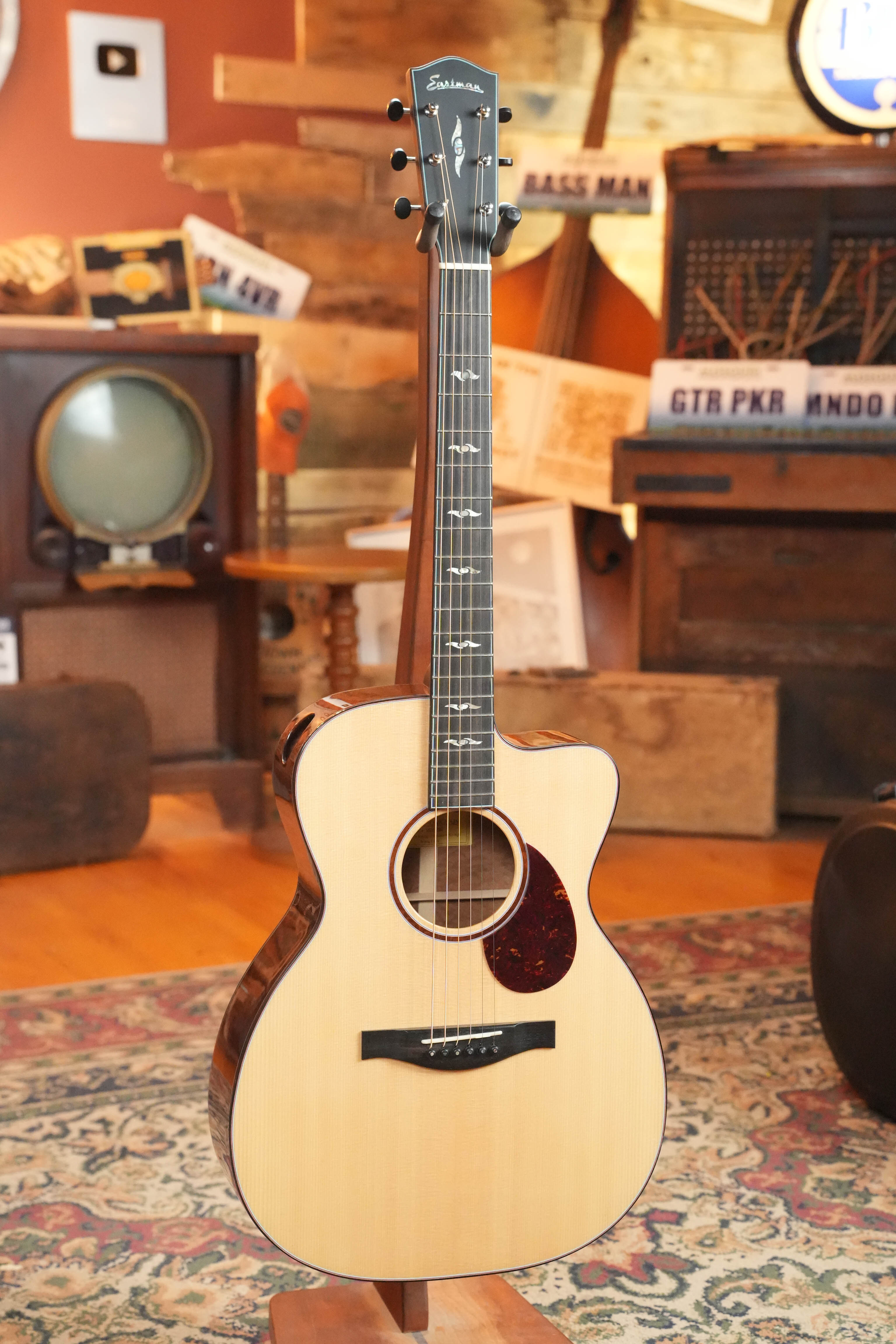 A *Dread-Not Certified Used* 2024 Eastman L-OMCE-QS Solid Engelmann Spruce/Quilted Sapele Orchestra Acoustic-Electric Guitar by Eastman Guitars stands upright on a patterned rug in a vintage-inspired room with musical decor.