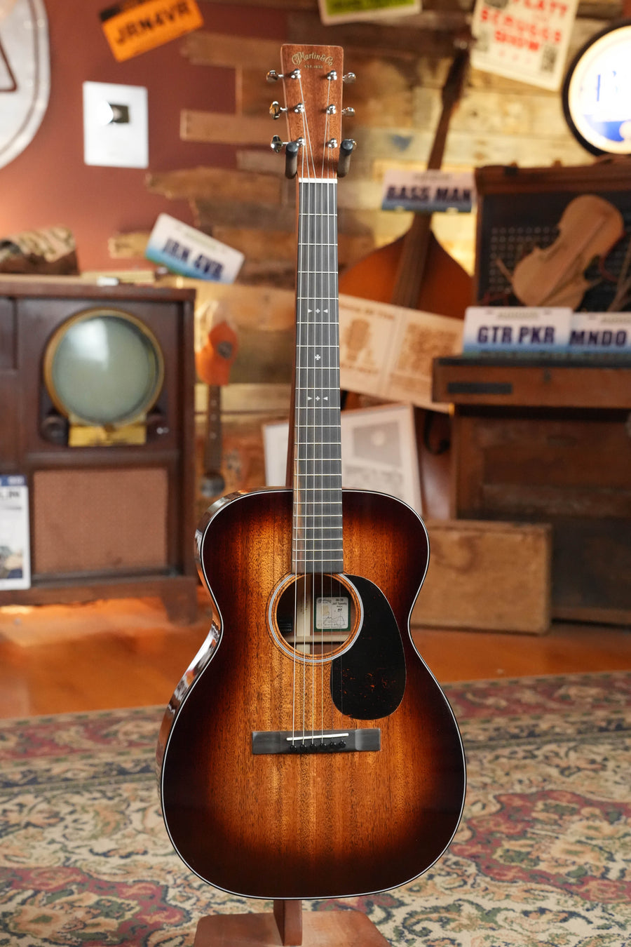 A *Dread-Not Certified Used* 2025 Martin OO-DB Jeff Tweedy All FSC Certified Mahogany Acoustic Guitar by Martin & Co. stands upright on a patterned rug in a cozy room with vintage decor, instruments, and wooden walls adorned with signs.