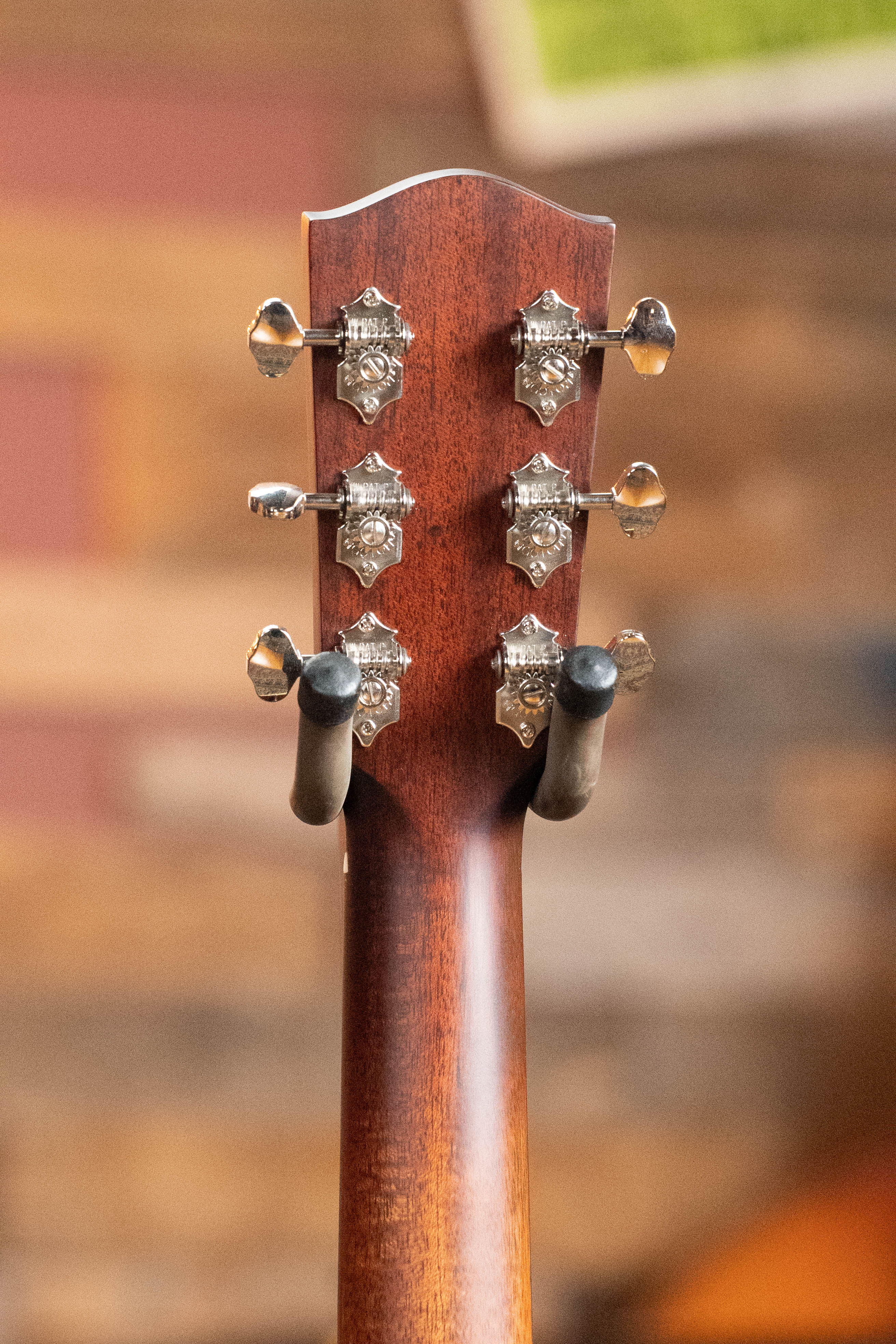 Close-up of the back of a *Dread-Not Certified Used* Eastman Guitars E6-LGSS-TC-SB acoustic guitar #9323, showing six silver tuners on a wooden neck as it rests on a wall-mounted holder against a blurred background.