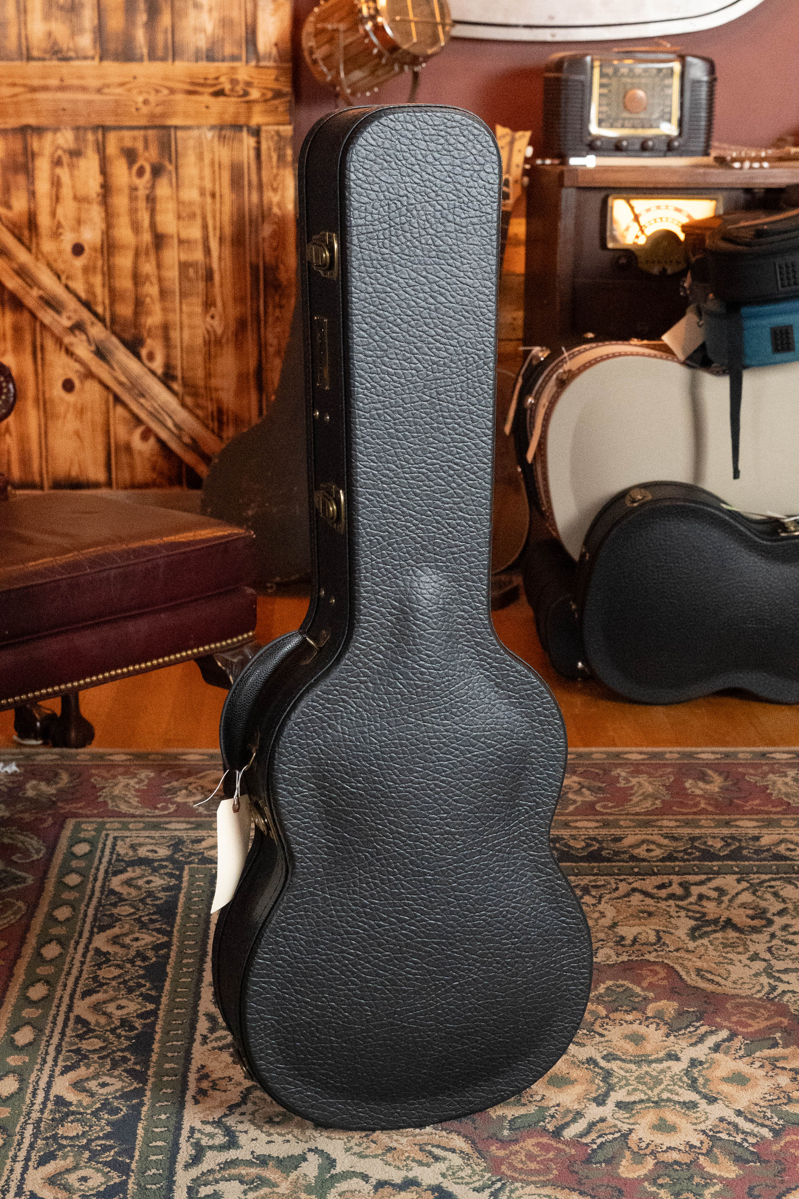 An Eastman Guitars *Dread-Not Certified Used* E6-LGSS-TC-SB Slope Shoulder Sunburst Acoustic Guitar with Sitka top and mahogany back/sides stands on a patterned rug in a cozy, vintage room beside a black hard-shell case.