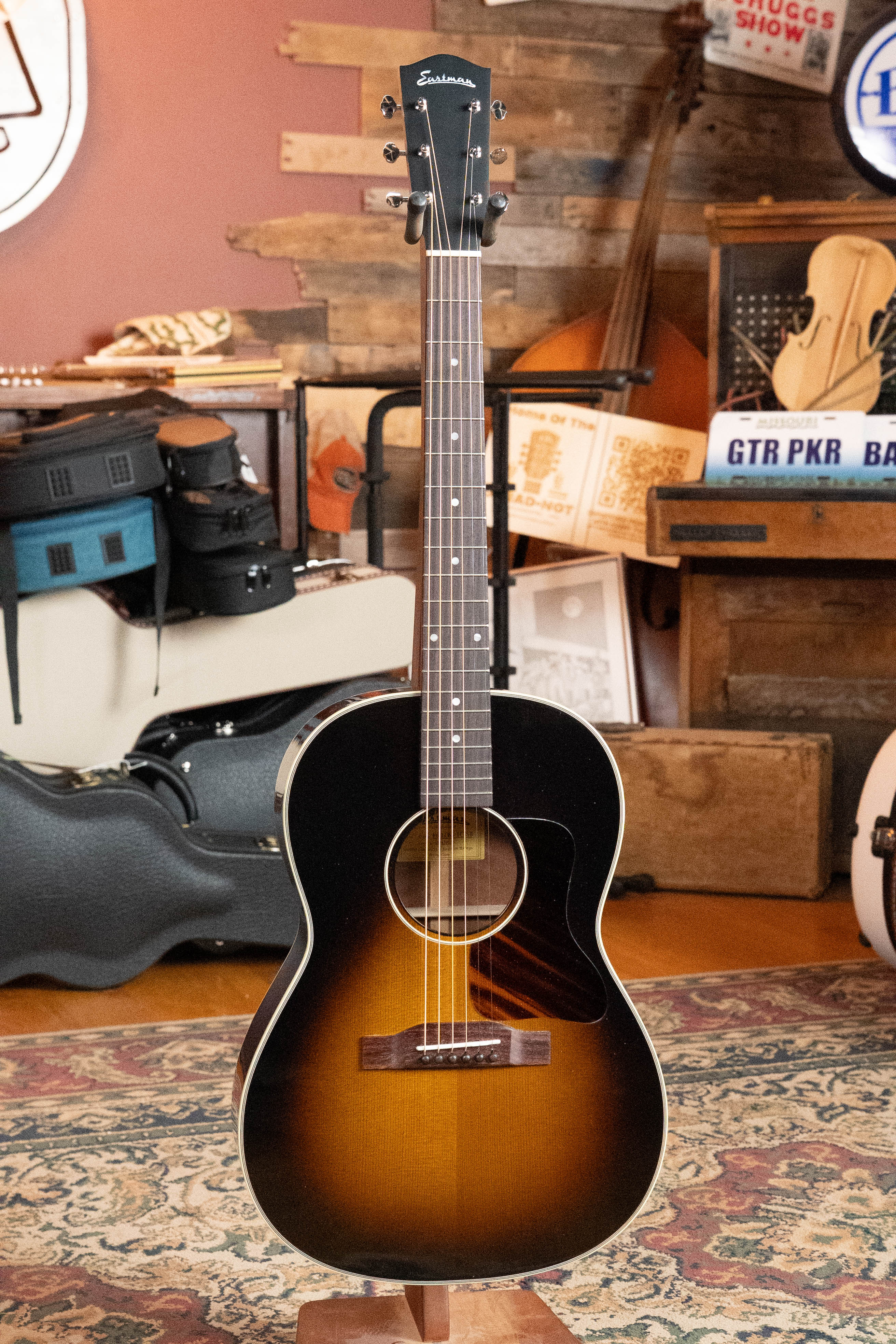 A *Dread-Not Certified Used* Eastman E6-LGSS-TC-SB slope-shoulder sunburst acoustic guitar with thermo-cured Sitka top and mahogany back/sides stands upright in a cozy, vintage-decor room filled with instruments and cases.