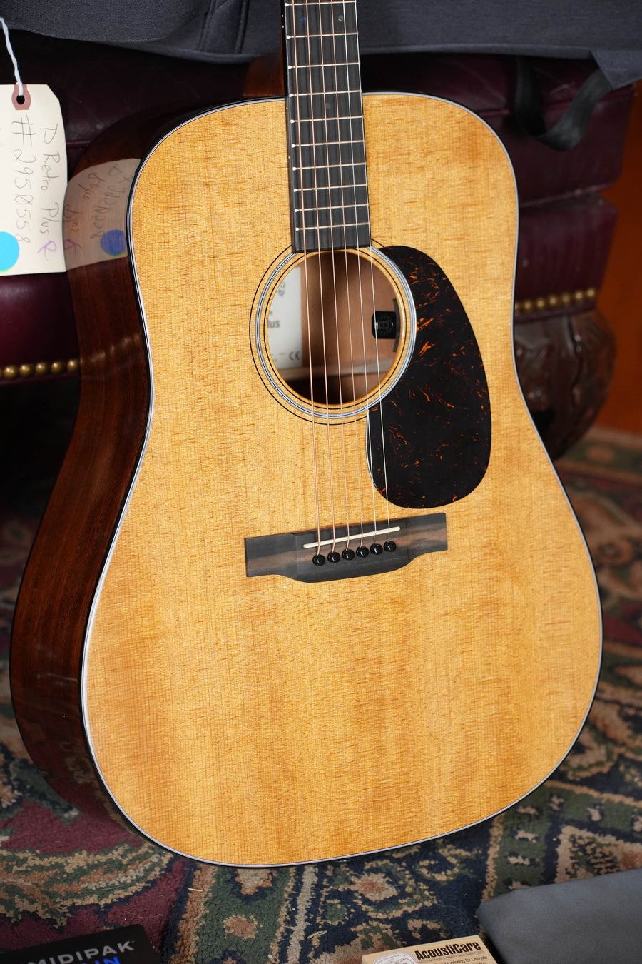 A close-up of a *Dread-Not Certified Used* Martin & Co. DE Retro Plus Dreadnought Guitar (#0558), with torrefied spruce top and mahogany, dark pickguard, strings, and handwritten tag, resting on a rug near a brown leather chair.