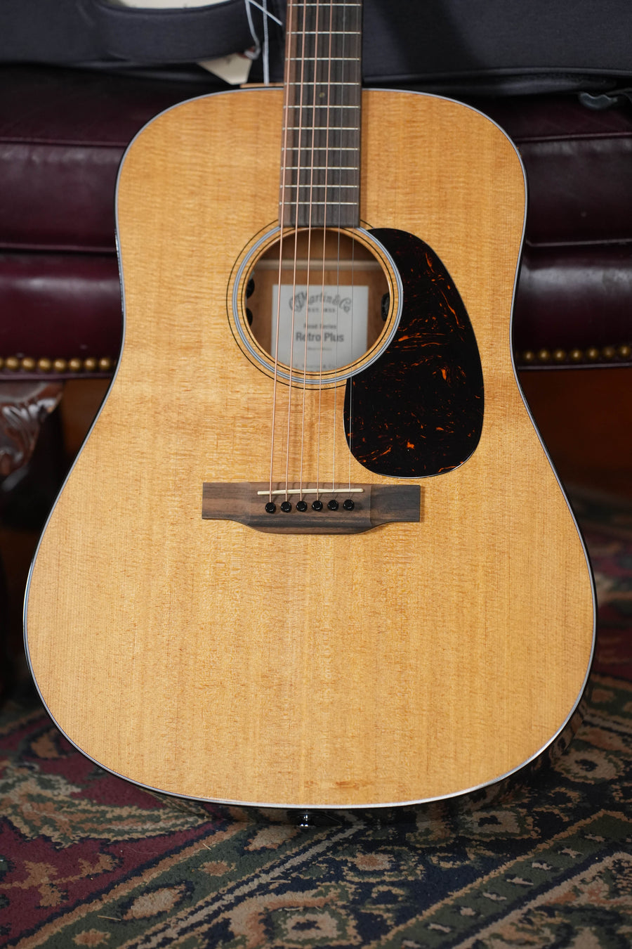 A close-up of a *Dread Not Certified Used* Martin & Co. DE Retro Plus Dreadnought Guitar with torrefied spruce top, mahogany back and sides, and Martin electronics, resting on maroon leather and a patterned carpet.