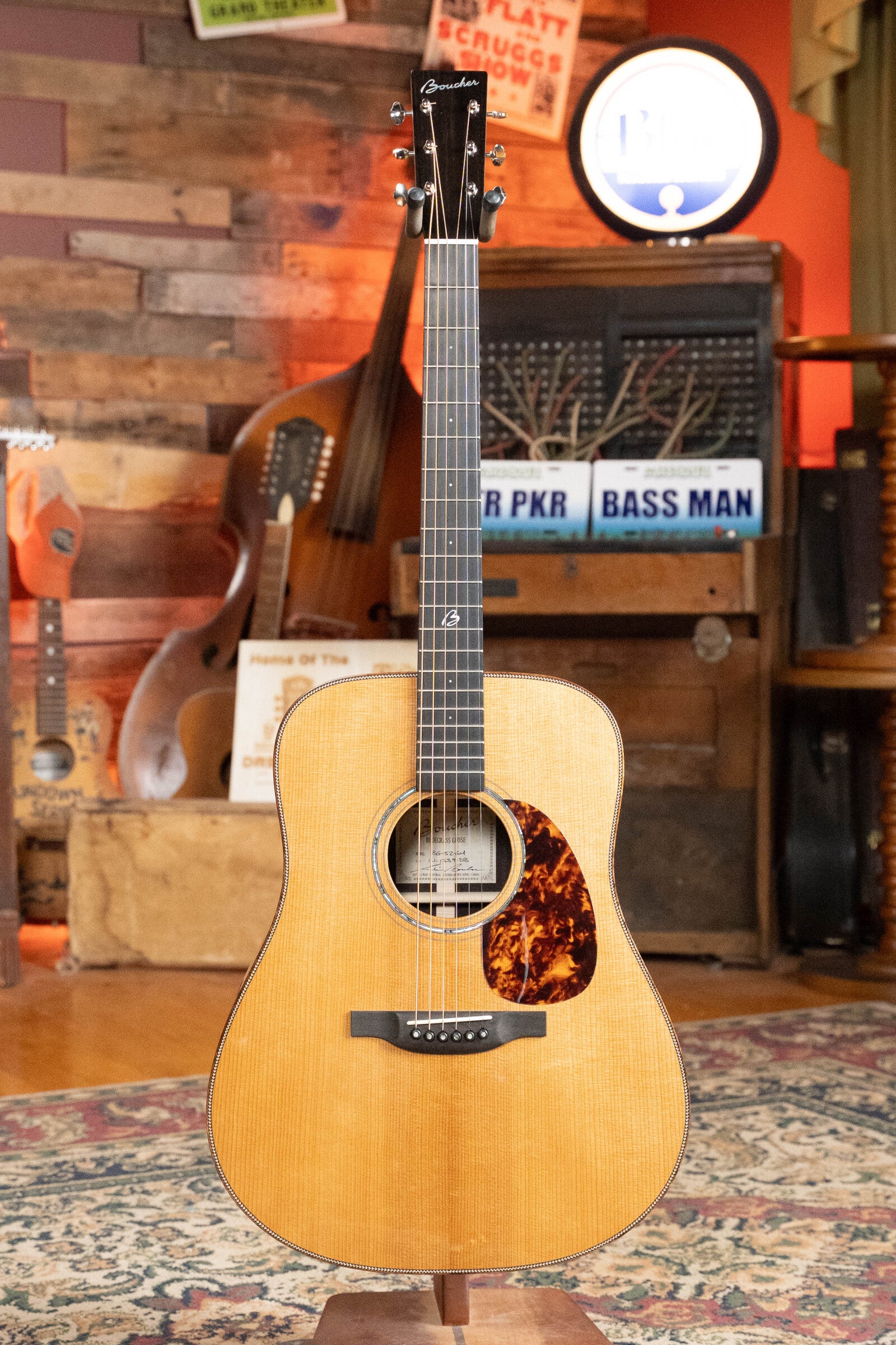 A *Dread-Not Certified Used* Boucher BG-52-GM Bluegrass Goose Series Dreadnought Acoustic Guitar stands on display in a cozy, music-themed room. It features Master Grade Adirondack Red Spruce and East Indian Rosewood.