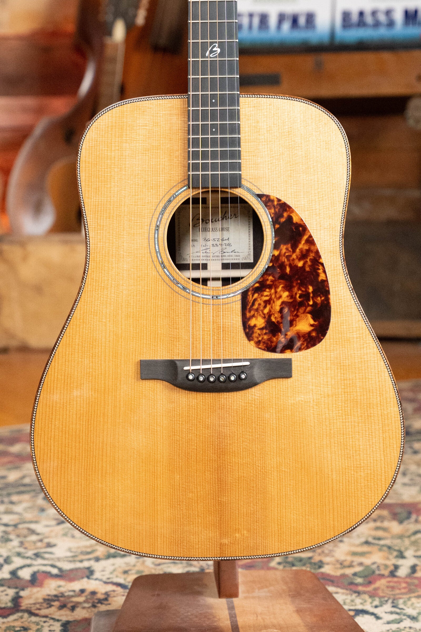 A close-up of a Boucher Guitars *Dread-Not Certified Used* BG-52-GM Bluegrass Goose Dreadnought (#1339), crafted from torrefied Adirondack Red Spruce and East Indian Rosewood, with a natural wood finish, on a patterned rug with blurred books and instruments.