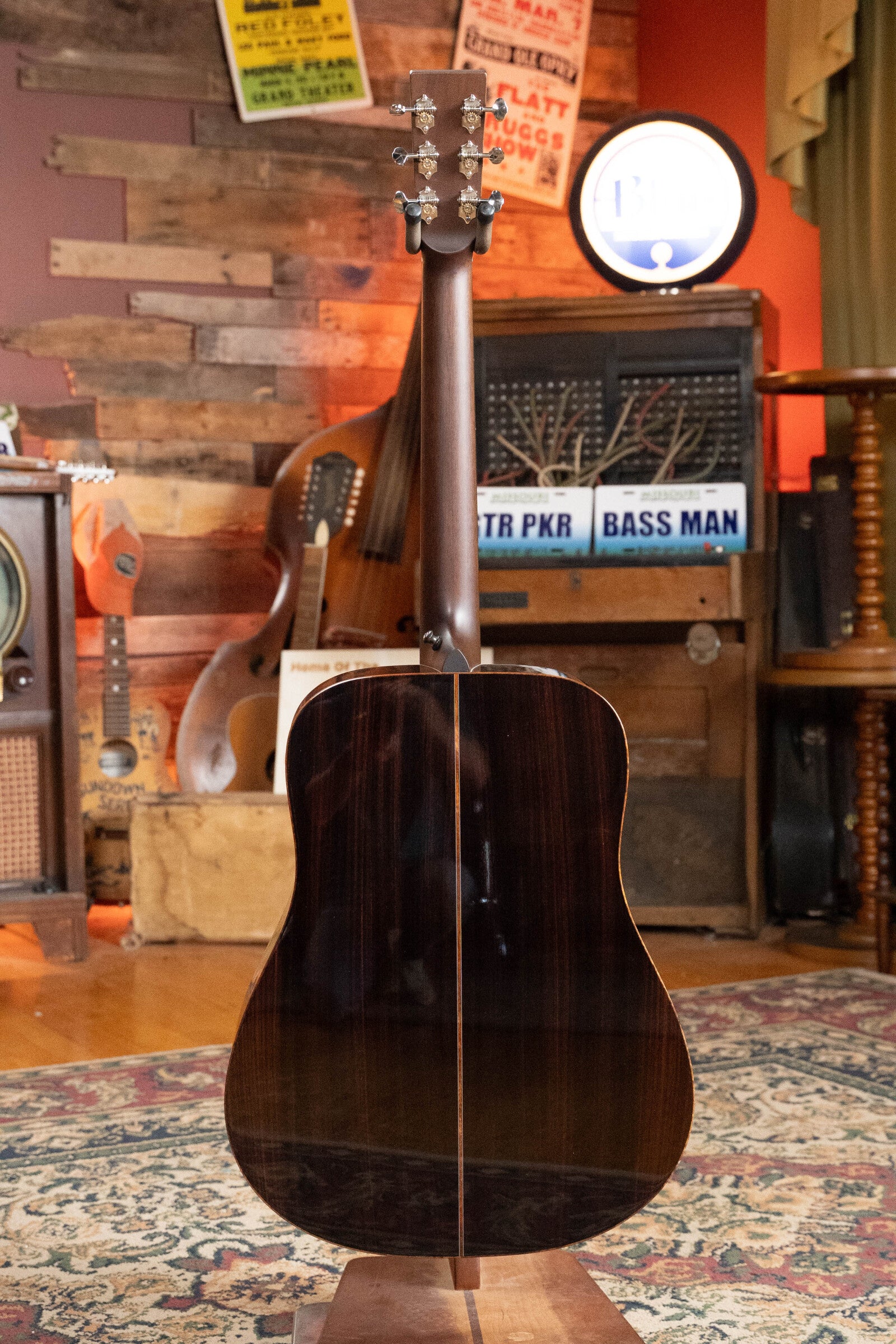 A *Dread-Not Certified Used* Boucher BG-52-GM Bluegrass Goose Series Dreadnought by Boucher Guitars, featuring a torrefied Adirondack spruce top and Indian Rosewood back, stands on a patterned rug in a cozy room with vintage decor and musical instruments.