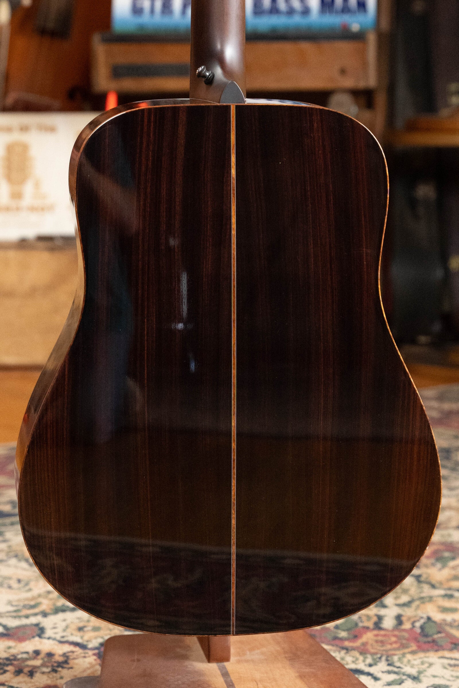 The image shows the back of a *Dread-Not Certified Used* Boucher BG-52-GM Bluegrass Goose Series Dreadnought by Boucher Guitars, featuring polished East Indian Rosewood with a thin center inlay, displayed on a stand in a vintage setting.