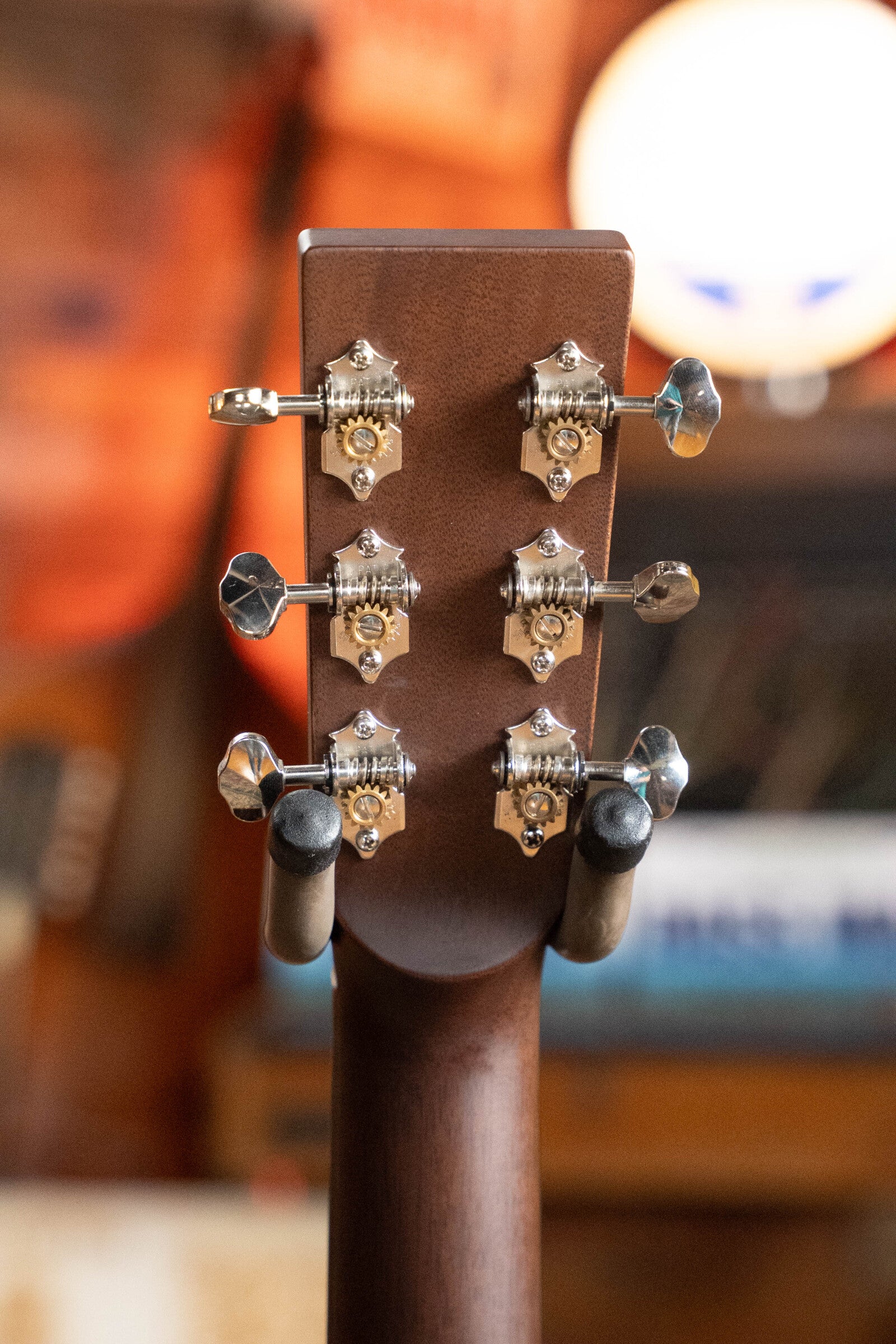 Close-up of the back of a *Dread-Not Certified Used* Boucher BG-52-GM Bluegrass Goose Series dreadnought headstock with six silver tuners, guitar on stand. Warm-toned, blurred background with circular light. Brand: Boucher Guitars.