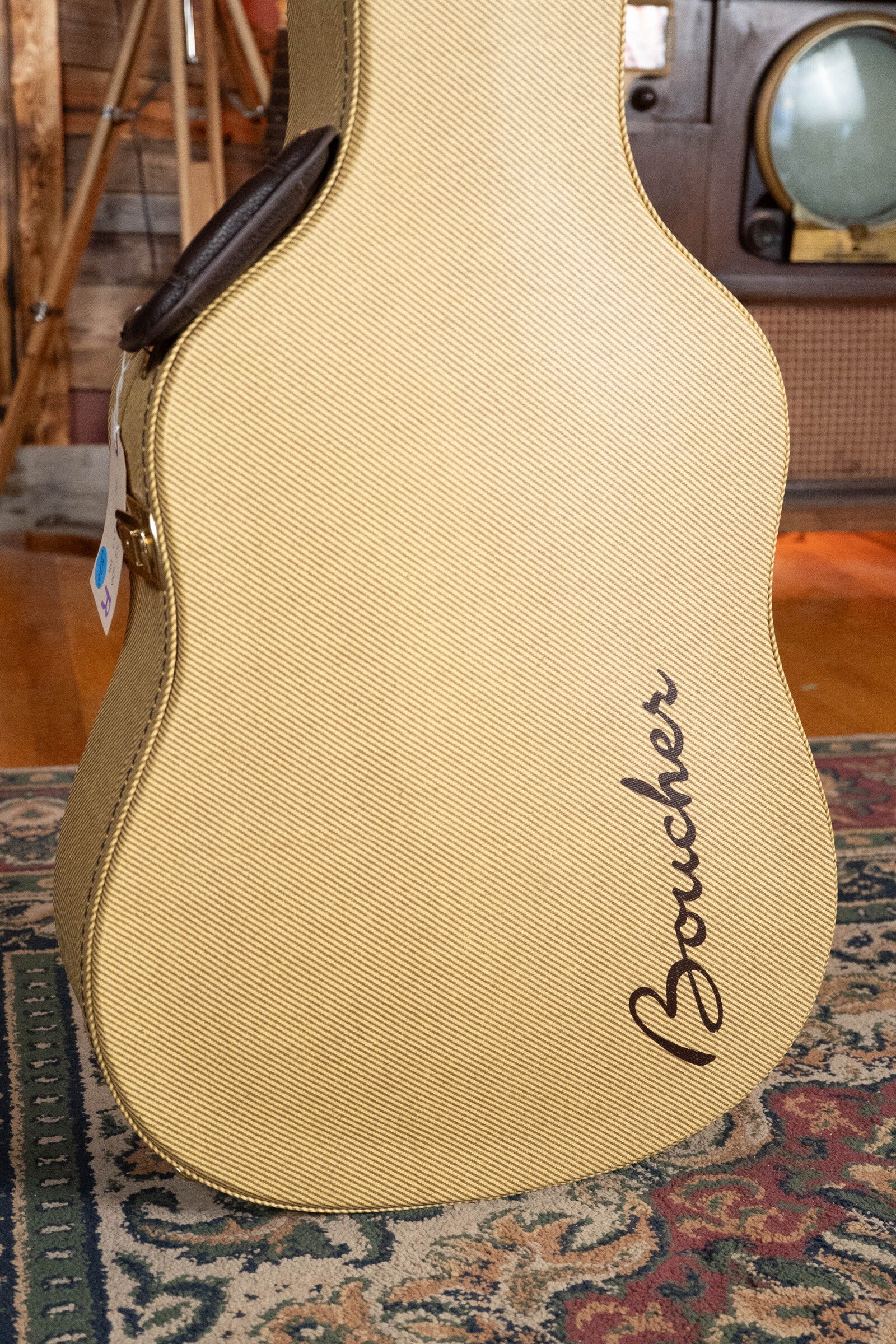 A tan tweed Boucher Guitars case rests on a patterned rug, with vintage-style furniture and a TV behind—suggesting the warm tones of the *Dread-Not Certified Used* BG-52-GM East Indian Rosewood Bluegrass Goose Series Dreadnought inside.