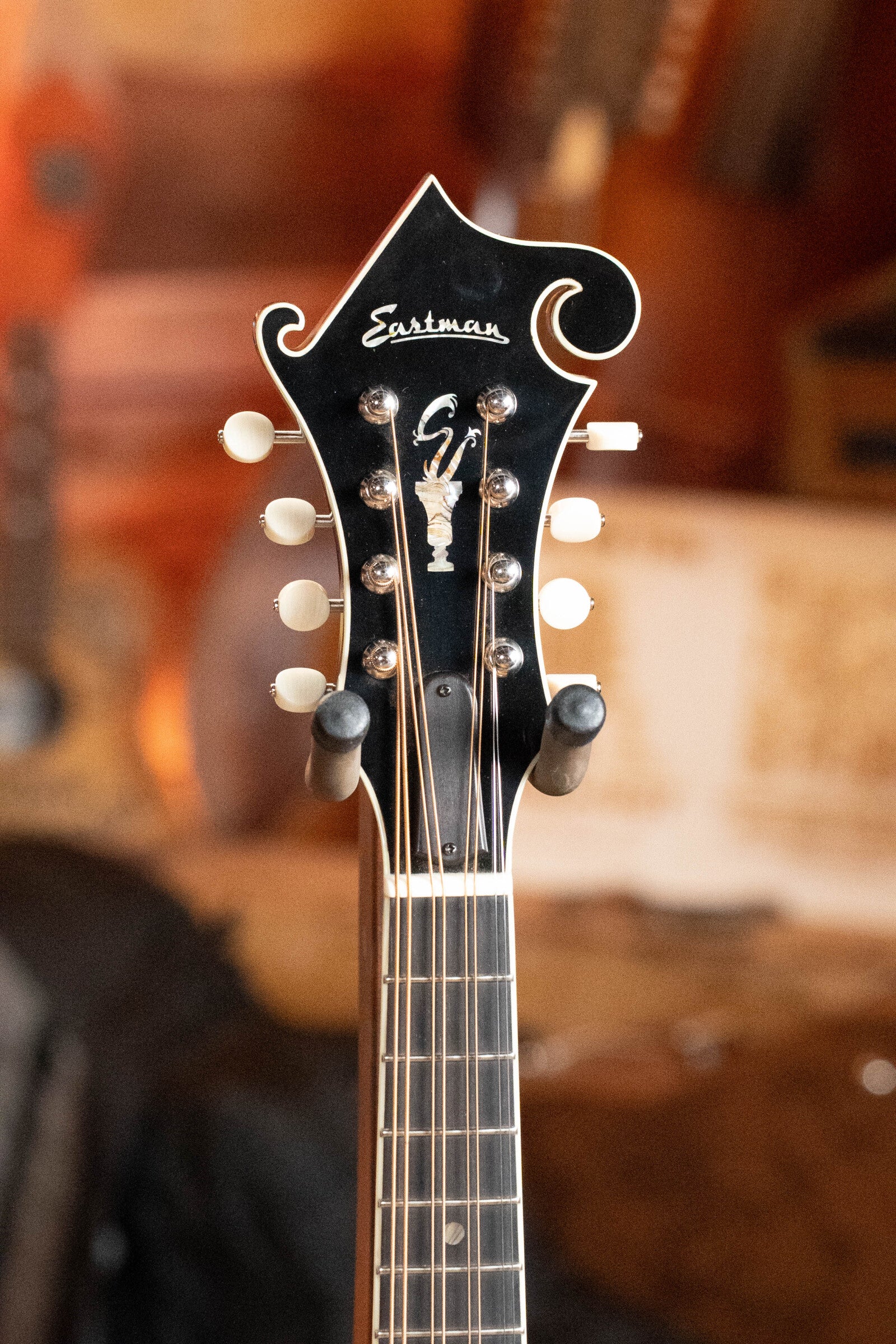 Close-up of the headstock on this *Dread-Not Certified Used* Eastman MDA815 F-Style Mandola Classic Finish #4978 by Eastman Guitars, showing tuning pegs, strings, and inlays with a blurred warm-toned background. Solid spruce top for rich tone.
