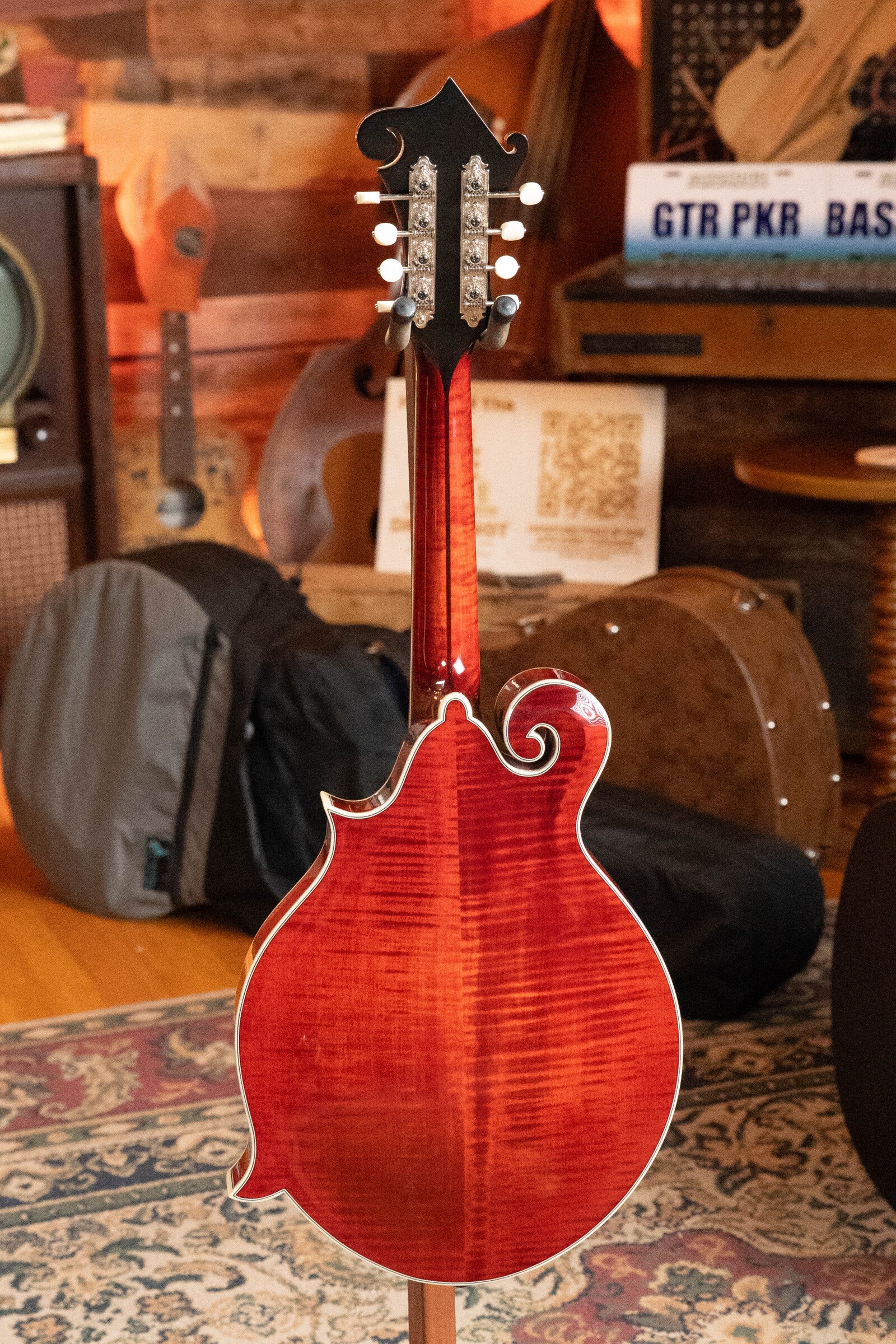 A *Dread-Not Certified Used* Eastman MDA815 F-Style Mandola Classic Finish #4978 by Eastman Guitars is displayed from the back, standing on a patterned rug in a cozy room with musical instruments and cases in the background.