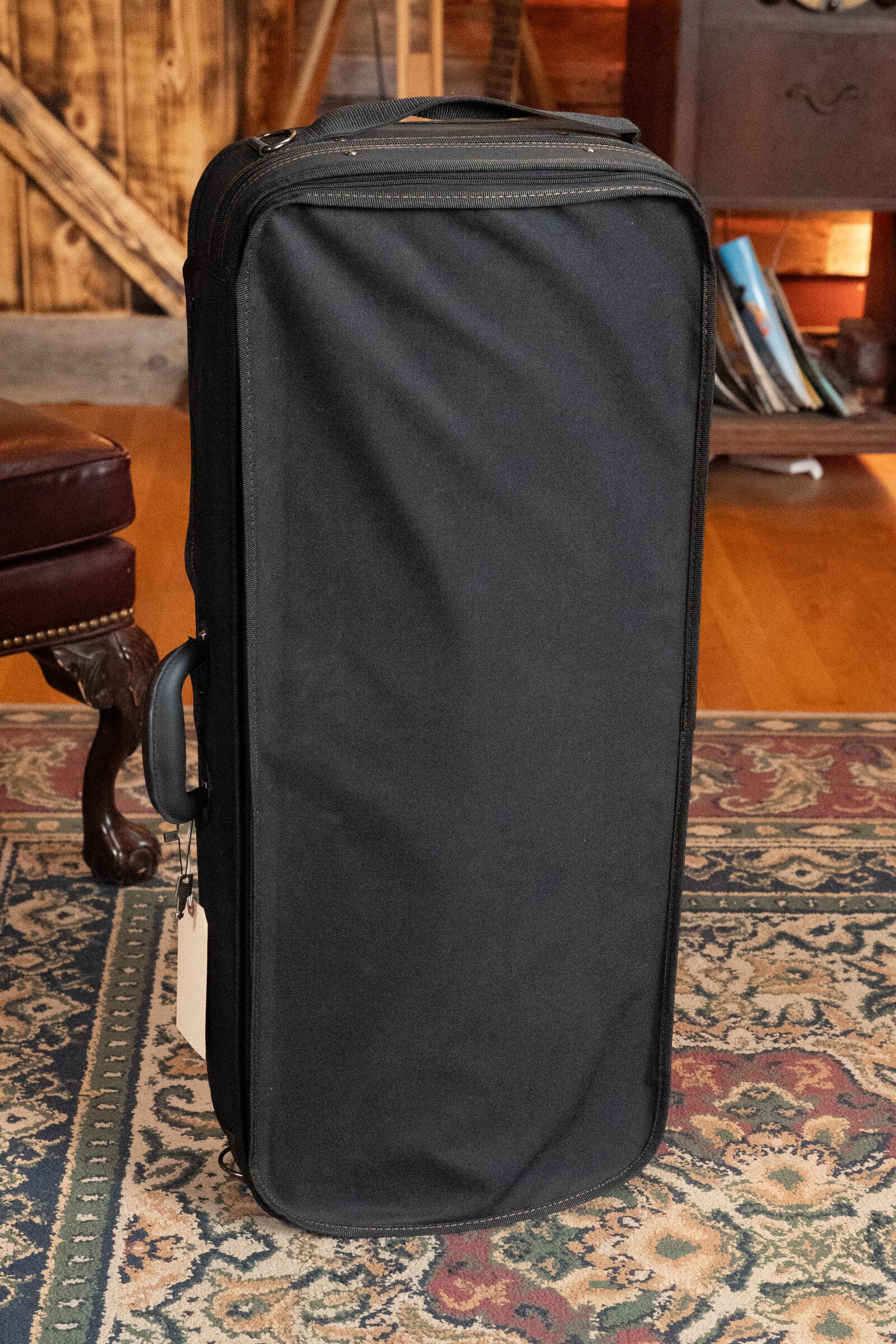 A black rectangular case with a handle stands on a patterned rug in a wood-paneled room, ideal for securely transporting your *Dread-Not Certified Used* Eastman MDA815 F-Style Mandola Classic Finish #4978 by Eastman Guitars.