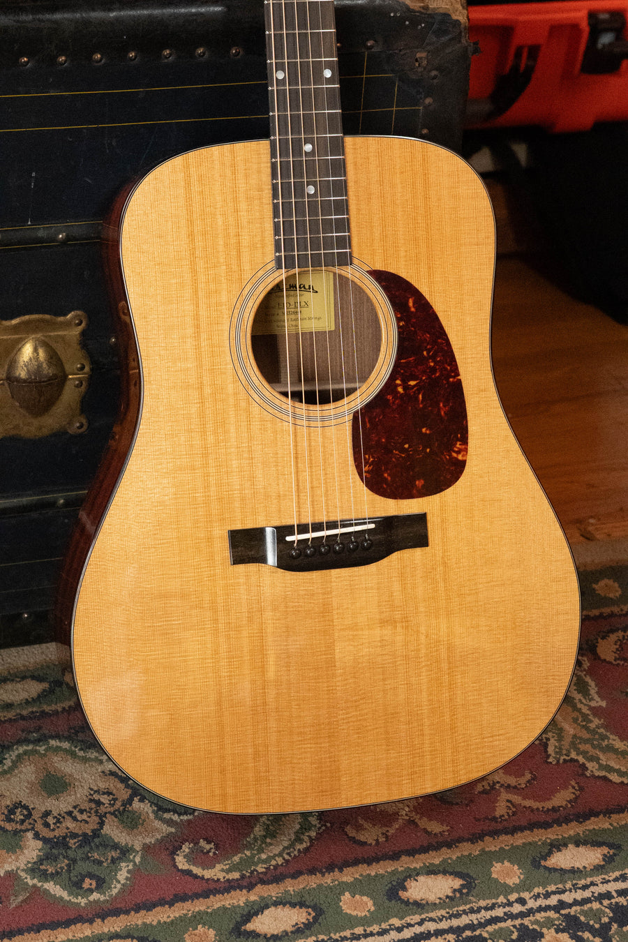A close-up of an Eastman Guitars E1D-DLX Sitka/Sapele Dreadnought Guitar w/Fishman Pickup #6444, featuring a natural finish and tortoiseshell pickguard, propped against a vintage trunk on a patterned rug.