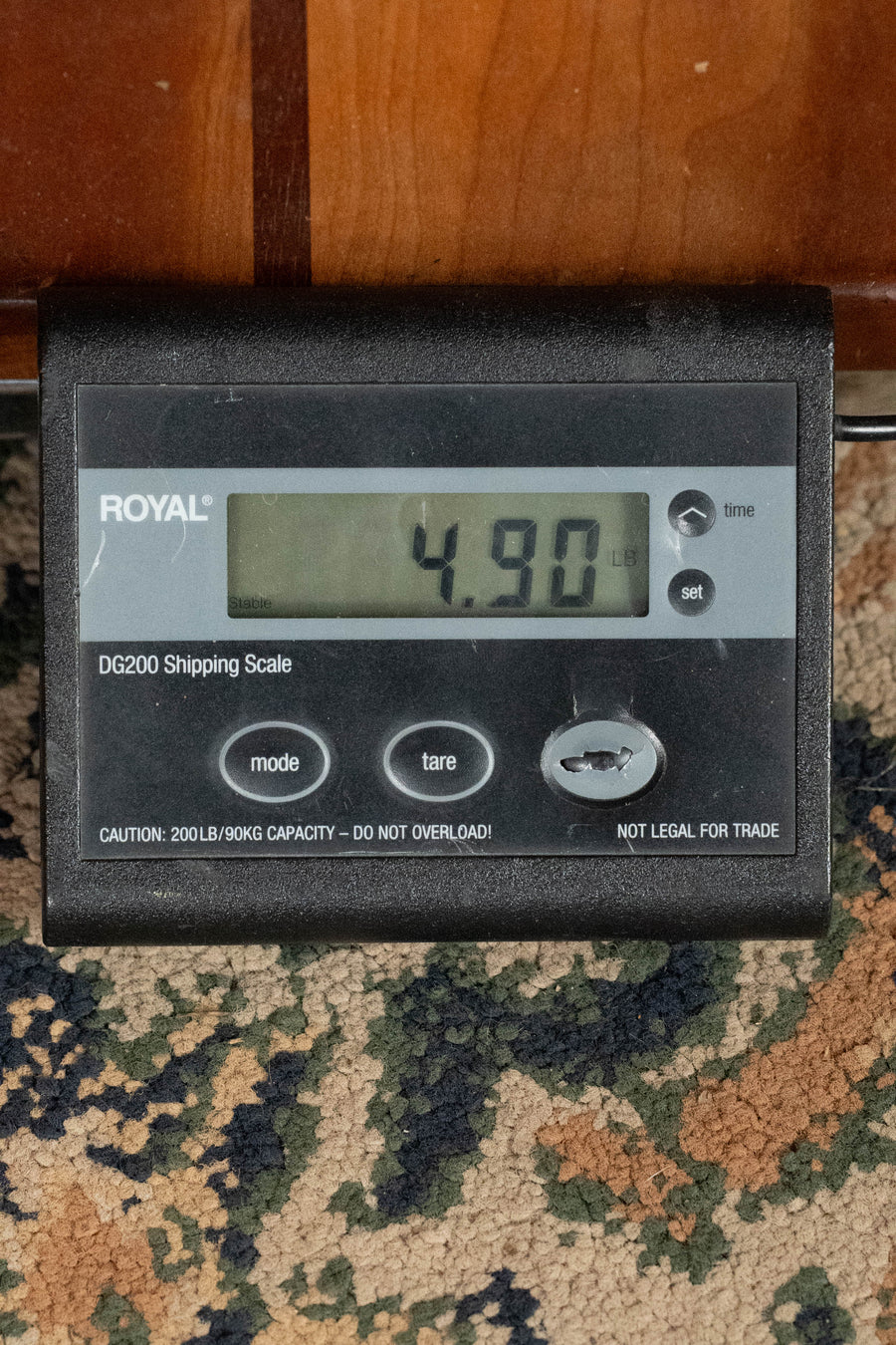 A digital shipping scale shows 4.90 pounds, possibly weighing an Eastman Guitars E1D-DLX Sitka/Sapele Dreadnought Guitar w/Fishman Pickup #6444. The scale is on a patterned carpet, with a wooden surface above it.