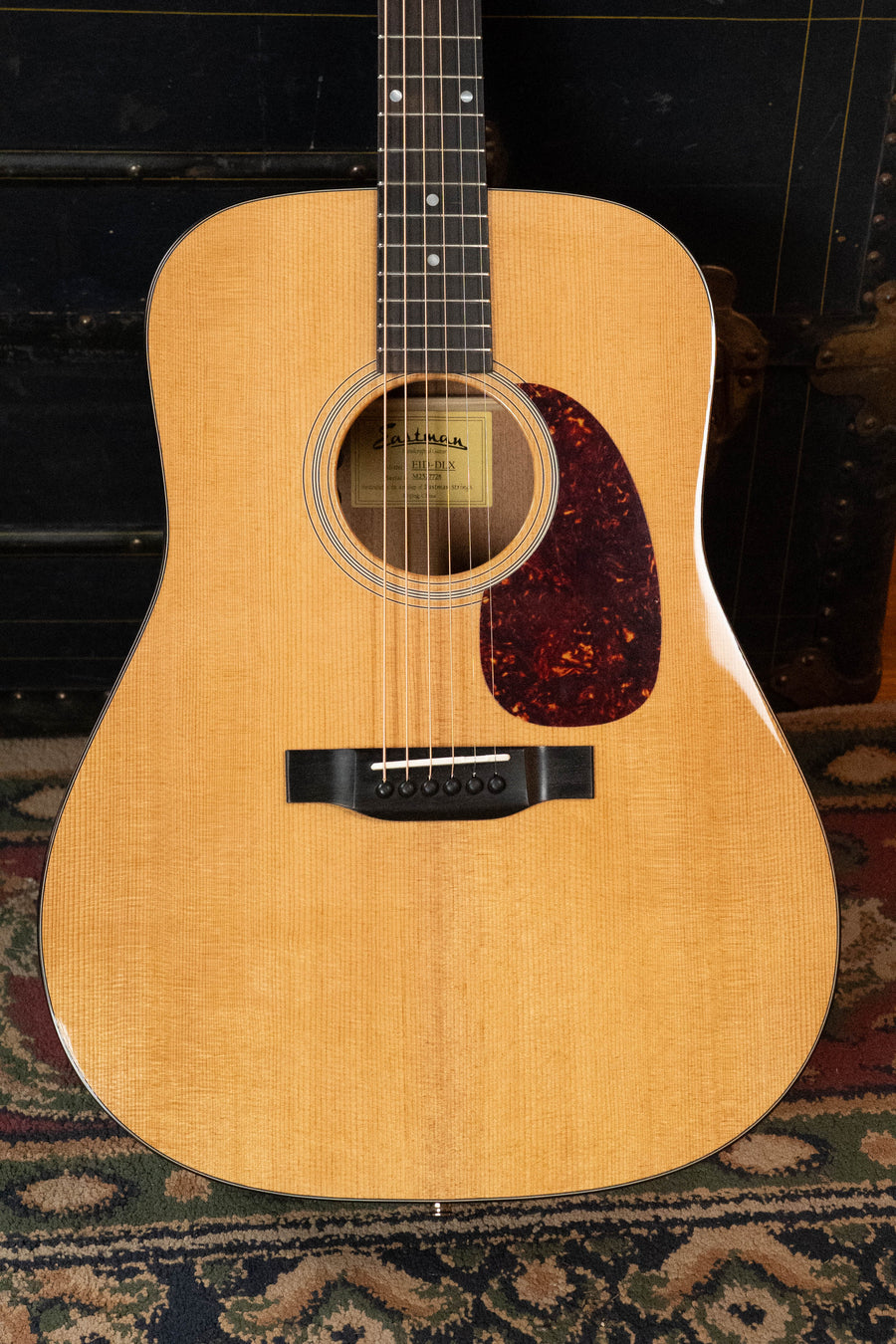A close-up of the Eastman Guitars E1D-DLX Sitka/Sapele Dreadnought Guitar w/Fishman Pickup #7728, featuring a natural finish, tortoiseshell-style pickguard, and black bridge, rests on a patterned rug against a dark background.