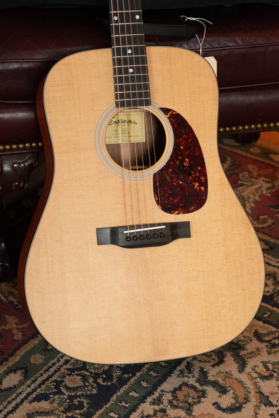 An Eastman Guitars E1D Sitka/Sapele Dreadnought Acoustic Guitar #7792 with a tortoiseshell pickguard rests on a patterned carpet in front of a dark red leather couch with brass stud accents.