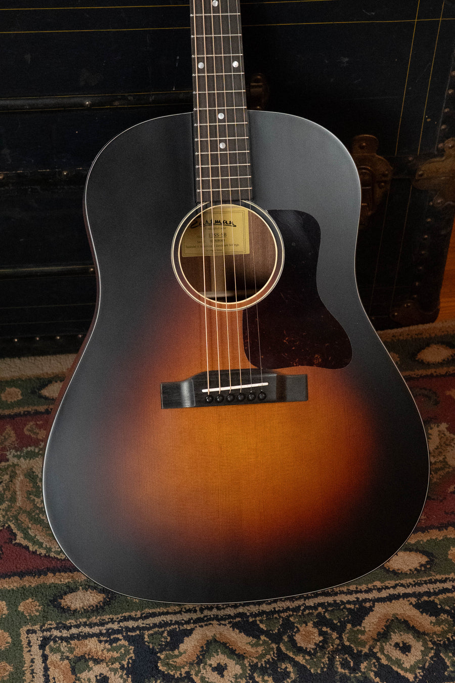 The Eastman Guitars E1SS-SB Sitka/Sapele Sunburst Slope Shoulder Dreadnought Acoustic Guitar #8068 stands upright against a dark chest, its glossy finish and pickguard gleaming above a patterned rug, with the sound hole label adding character.
