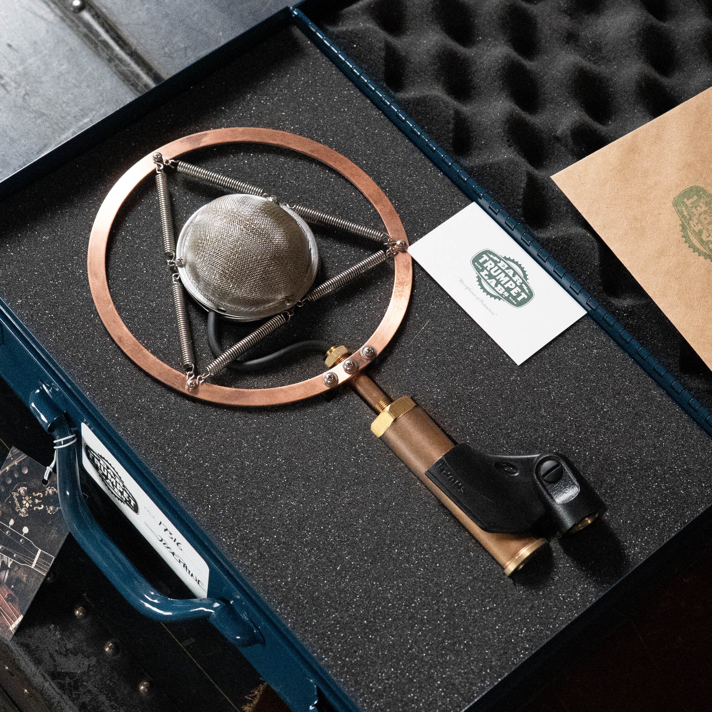 The Ear Trumpet Labs Josephine Microphone, a vintage-style large-diaphragm condenser with a copper and brass frame, rests in a foam-lined blue case beside a brown envelope and a “Plosive Planet” label card.