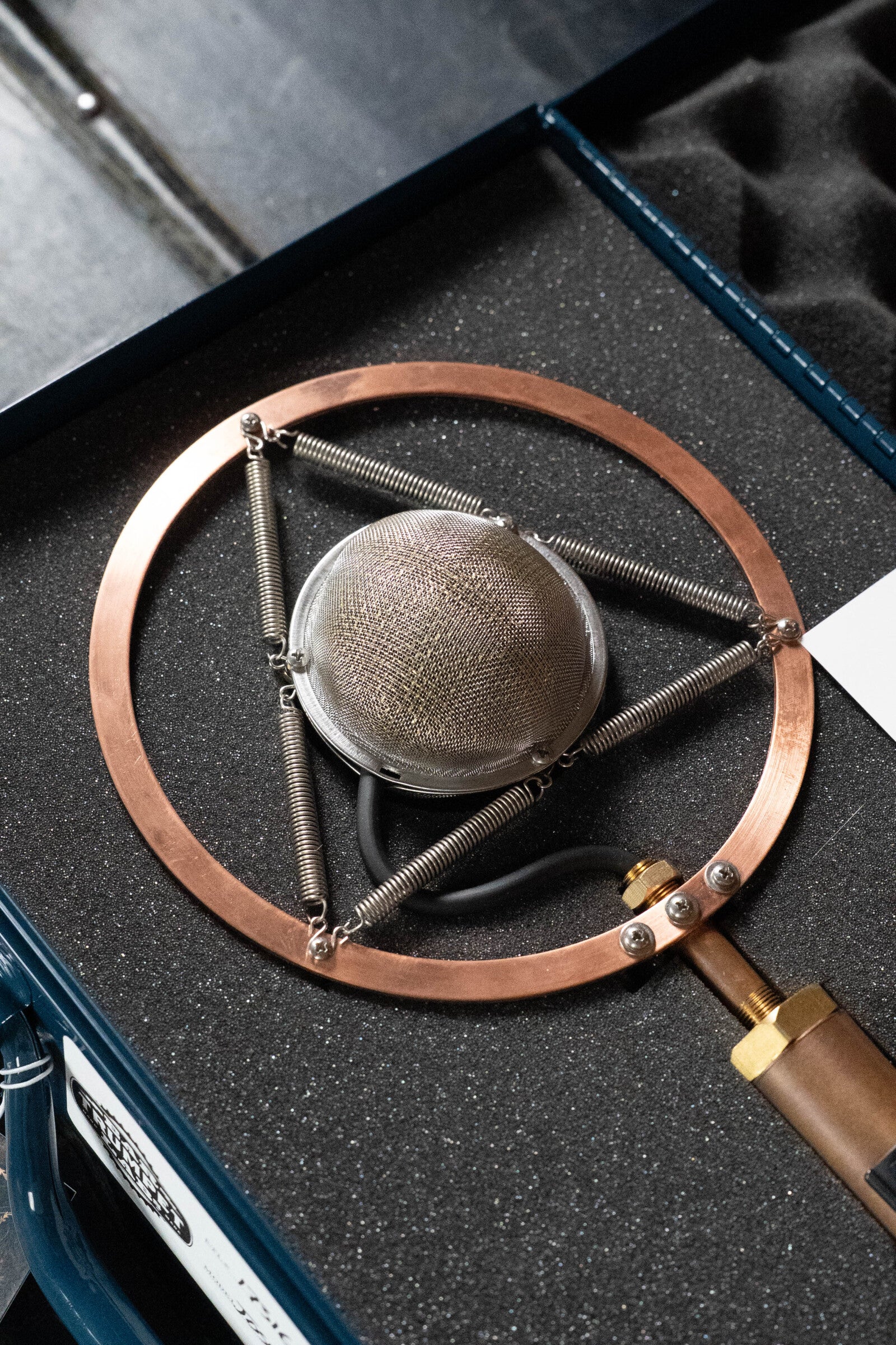 A close-up of the Ear Trumpet Labs Josephine Microphone, featuring a copper ring, metal springs, and mesh grille, resting in its foam-lined carrying case.