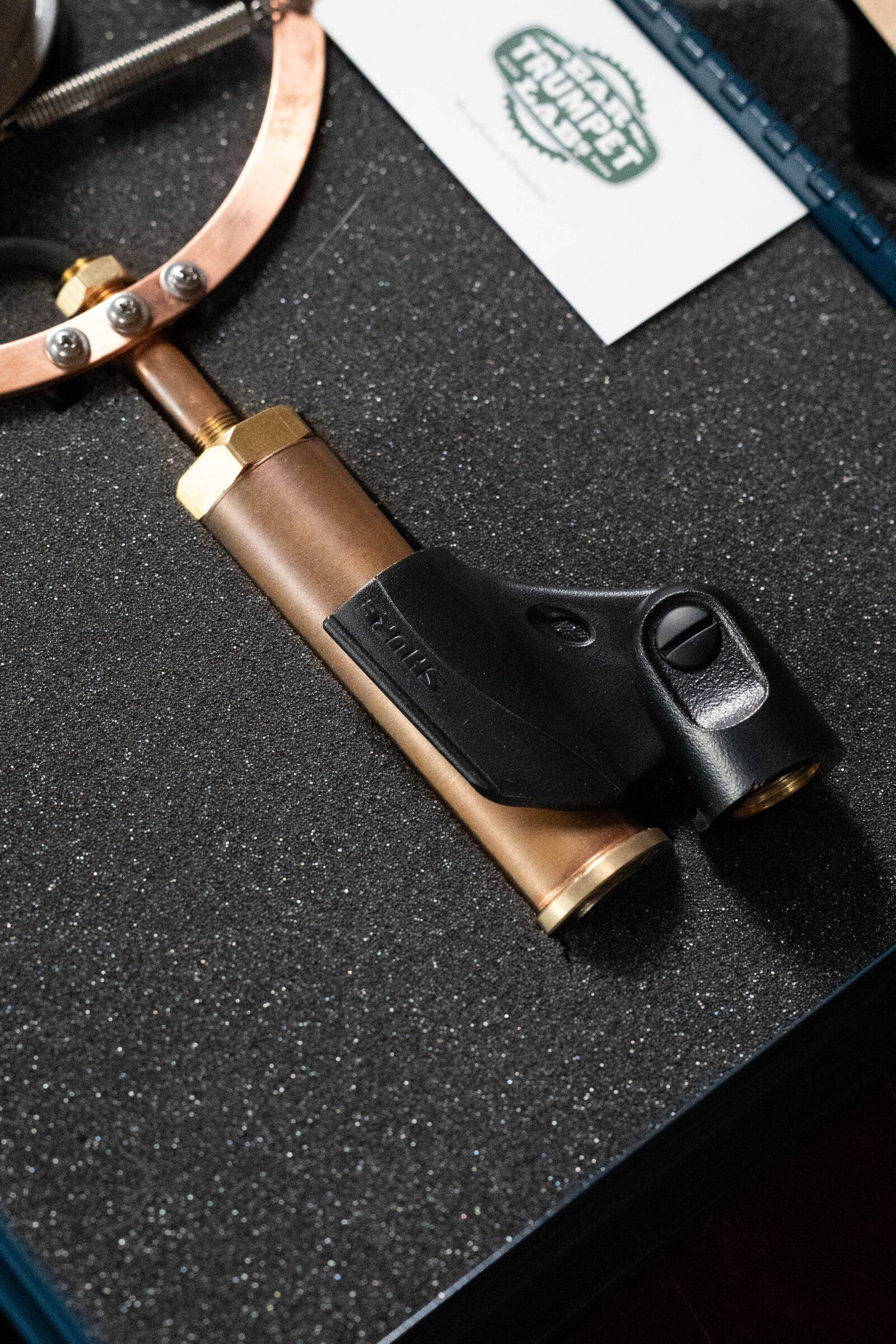 Close-up of an Ear Trumpet Labs Josephine large-diaphragm condenser microphone by Ear Trumpet Labs, featuring brass and black finish with a handle, resting in a foam-lined case. A white card with green text appears in the background.