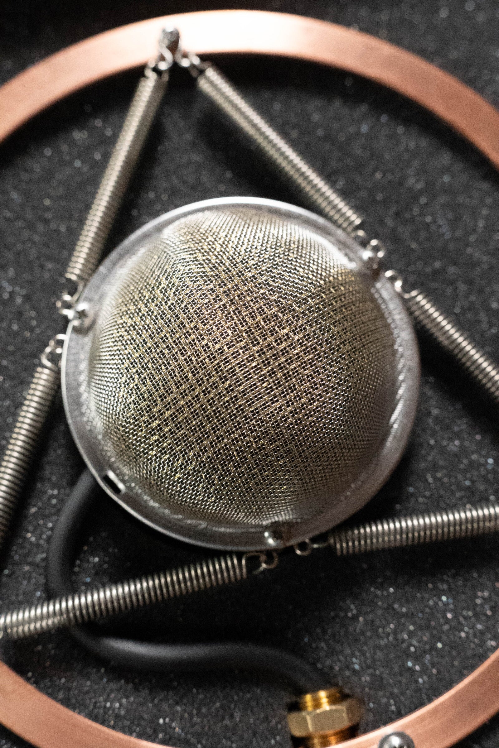 A close-up of the Ear Trumpet Labs Josephine Microphone shows its metal mesh dome secured by springs inside a copper frame on black foam, with a black wire and brass connector—features known for feedback resistance—visible at the bottom.