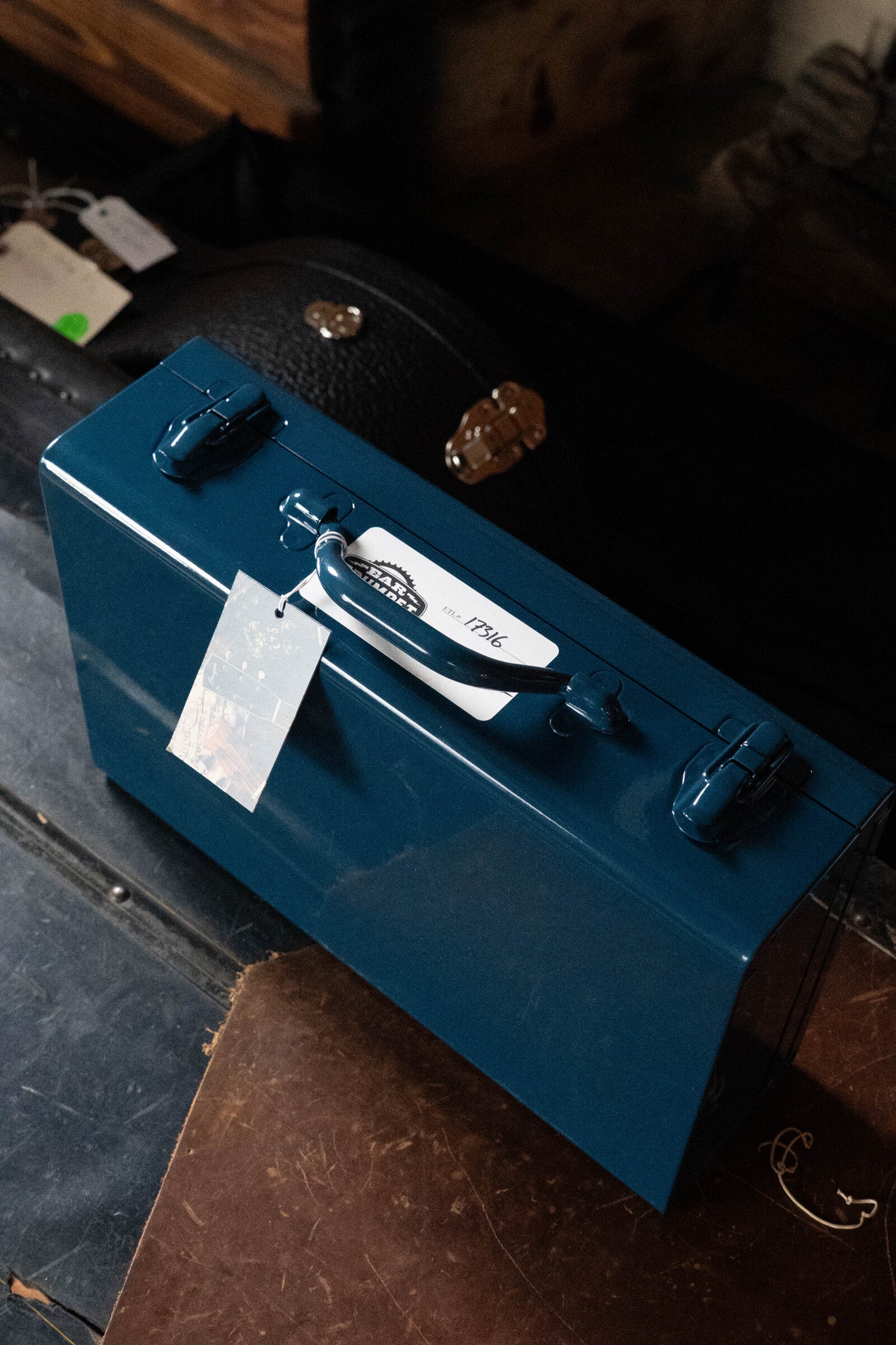 A blue metal briefcase with a handle and latches sits on a brown surface. A white tag labeled Ear Trumpet Labs is attached, indicating it stores the Ear Trumpet Labs Josephine Microphone. A black hard case is partially visible in the background.