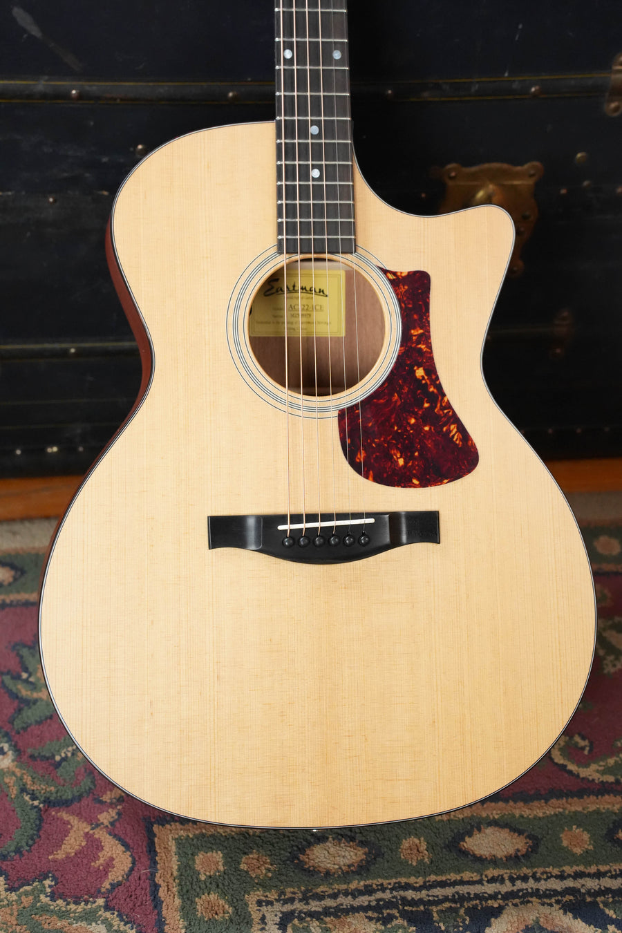 A close-up of an Eastman Guitars AC122-1CE Sitka/Sapele Grand Auditorium Cutaway Guitar w/Fishman Pickup rests upright on a patterned rug before a dark trunk with brass hardware. It features a natural finish, dark pickguard, and black bridge.