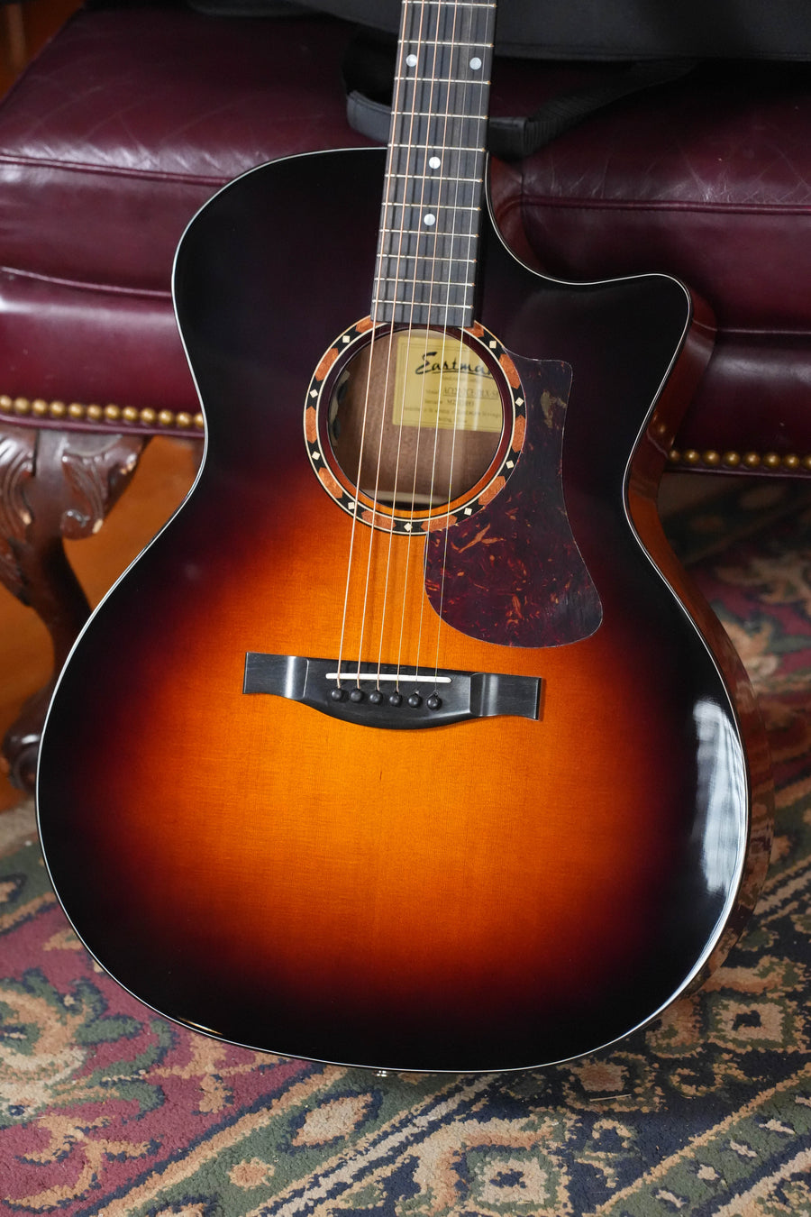 An Eastman AC122-2CE-DLX-SB Grand Auditorium Sunburst Cedar/Sapele Cutaway guitar with Fishman pickup rests on a patterned rug, leaning against a burgundy leather sofa with brass nailhead trim.