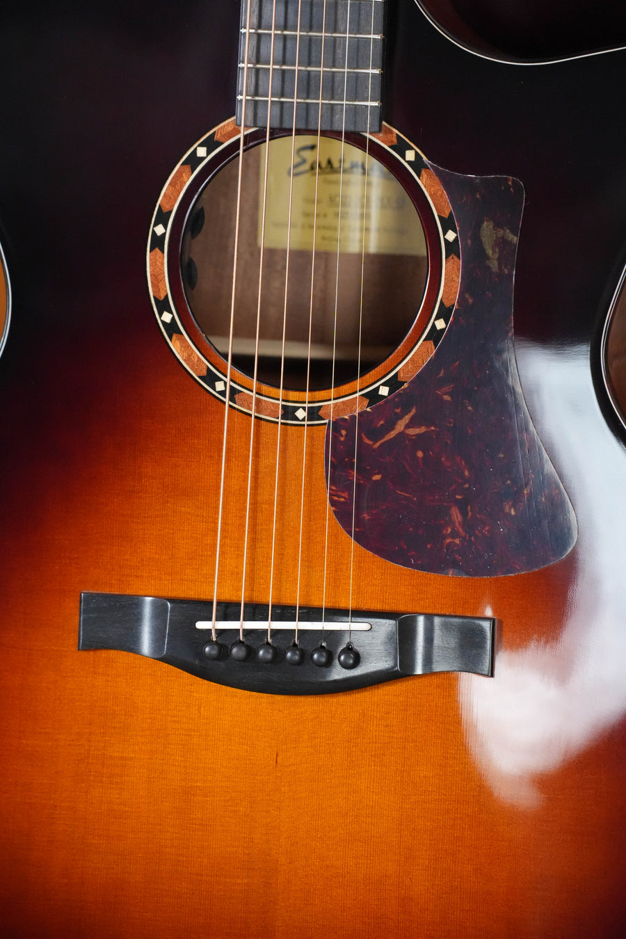 Close-up of the Eastman Guitars AC122-2CE-DLX-SB Grand Auditorium Guitar (#3693), highlighting six strings, dark bridge, decorated soundhole, tortoiseshell pickguard, glossy sunburst cedar top, and sapele back and sides.