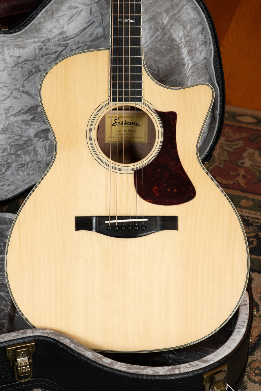 An Eastman Guitars AC322CE Sitka/Mahogany Cutaway Grand Auditorium Guitar w/LR Baggs Pickup (#3249) sits in an open hardshell case, showing its light body, dark pickguard, and strings on a patterned rug.
