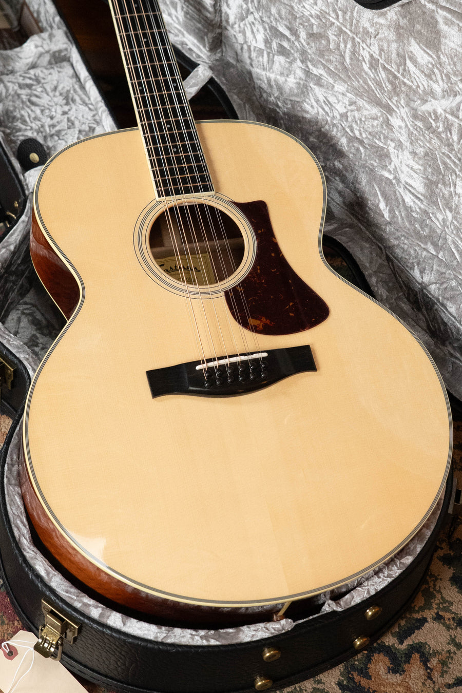 An Eastman Guitars AC330E-12 Sitka/Mahogany Jumbo 12-String with LR Baggs pickup (#5666) lies in an open padded case on a patterned rug, featuring a light Sitka spruce top and dark mahogany sides.