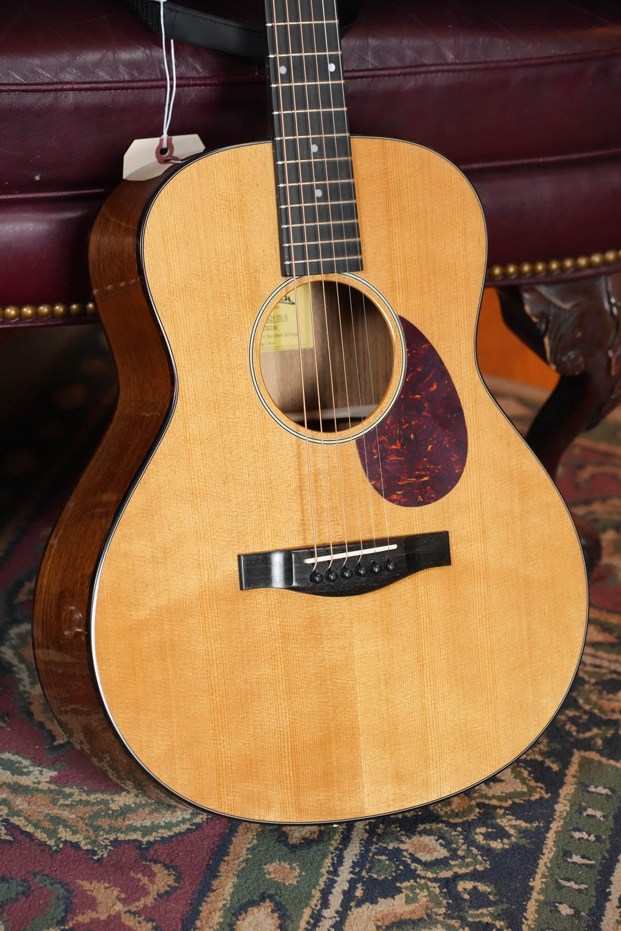 An Eastman Guitars ACTG2E-DLX Sitka/Ovangkol Travel Guitar w/Fishman Pickup #2361, featuring a light Sitka top, dark Ovangkol sides, and a tortoiseshell pickguard, rests against a maroon leather chair on a patterned rug.