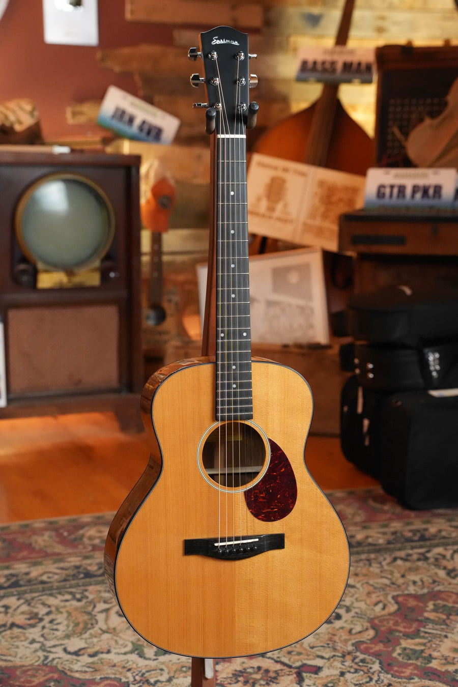 An Eastman Guitars ACTG2E-DLX Sitka/Ovangkol Travel Guitar w/Fishman Pickup (#2361) stands on a colorful rug in a cozy room with wood floors, vintage decor, retro TV, cases, and music signs—a perfect showcase for its tone and style.
