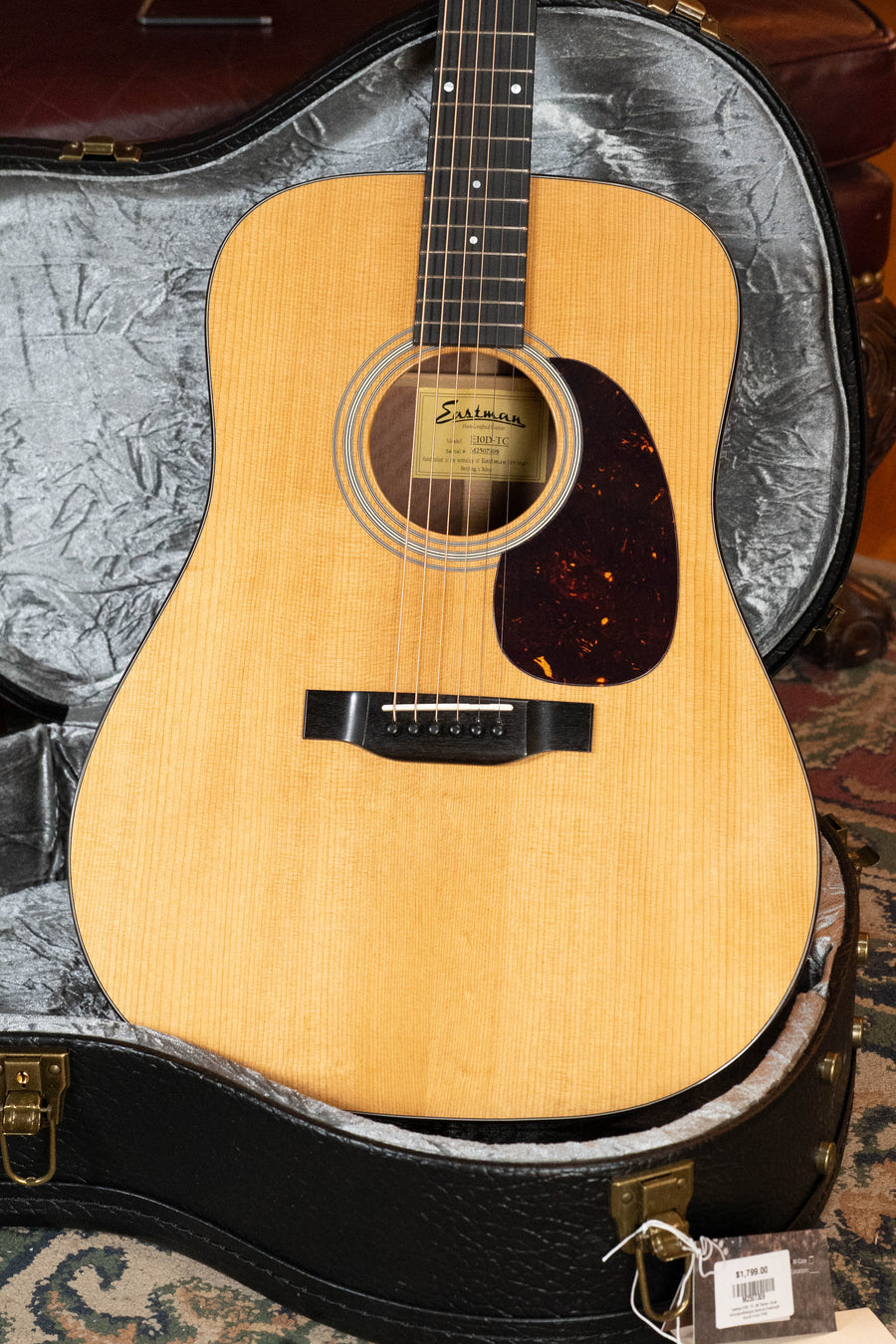 An Eastman E10D-TC Thermo-Cured Adirondack/Mahogany Dreadnought Acoustic Guitar by Eastman Guitars rests in an open black case with a tag on the latch, set atop a patterned rug—classic looks echoing vintage-inspired tone.