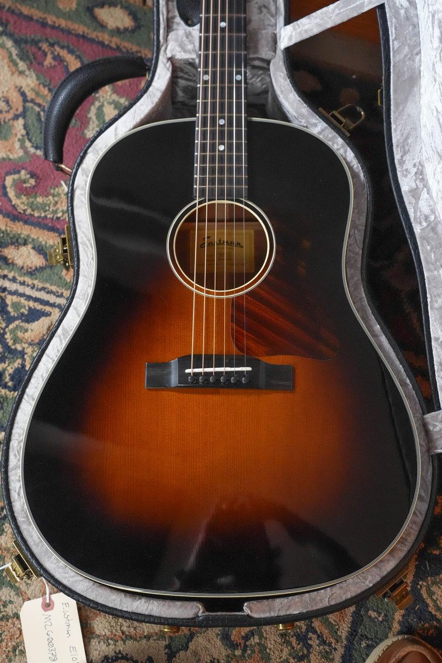 An Eastman Guitars E10SS-TC Thermo-Cured Adirondack/Mahogany Sunburst Slope Shoulder Dreadnought Acoustic Guitar #0378 sits in an open, padded case on a patterned rug, with a handwritten tag attached to the handle.
