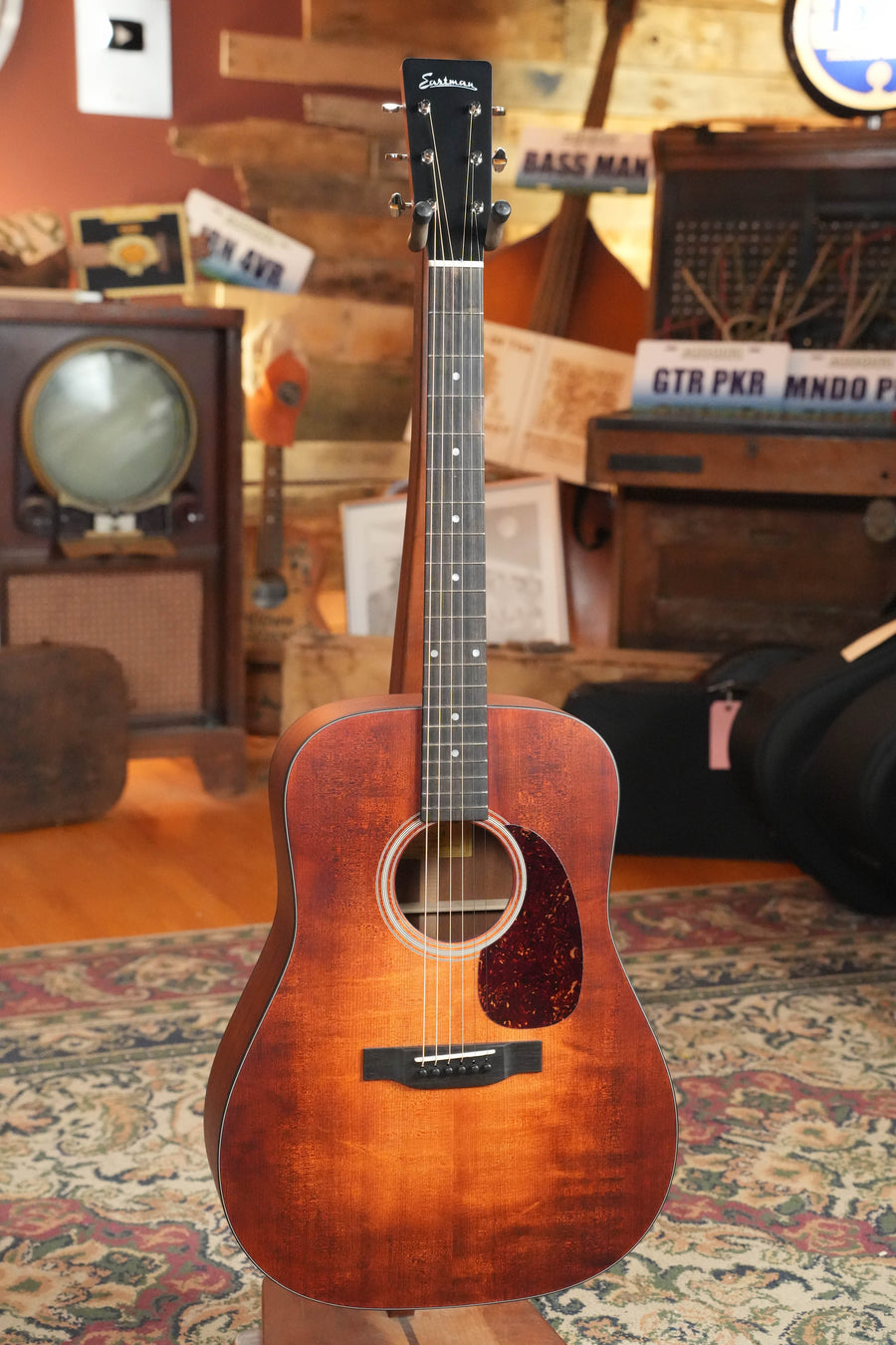 An Eastman E1D-CLA Classic Finish Dreadnought Acoustic Guitar by Eastman Guitars stands upright on a patterned rug in a cozy, wood-paneled room filled with vintage electronics and signs.